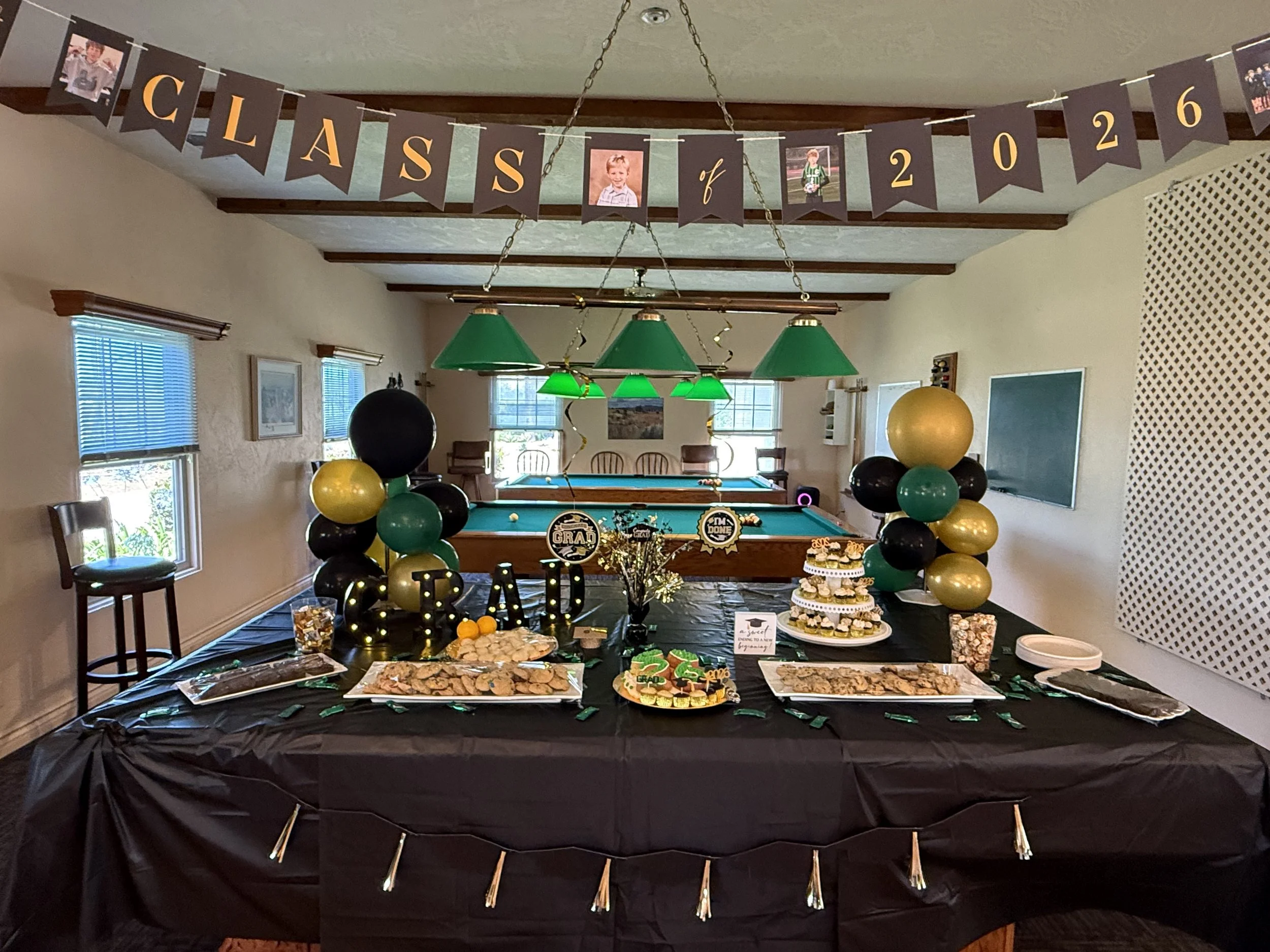 A party setup for a graduation celebration with a black tablecloth, cupcakes, cookies, and a variety of decorations including green, black, and gold balloons, a large sign that says 'GRAD,' and a banner with photos and words 'Class of 2026' hanging from the ceiling.