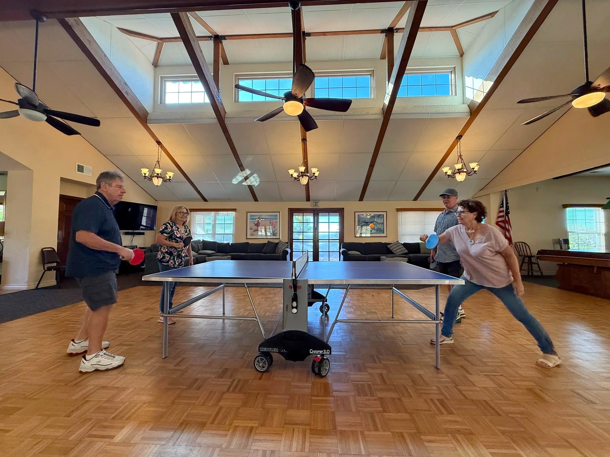 Four people are playing ping pong in a room with a wooden floor and large windows. Two women and two men are engaged in the game, one woman is holding a paddle and a ball, and the others are watching or preparing to play. The room has ceiling fans and chandeliers, with artwork and an American flag visible in the background.