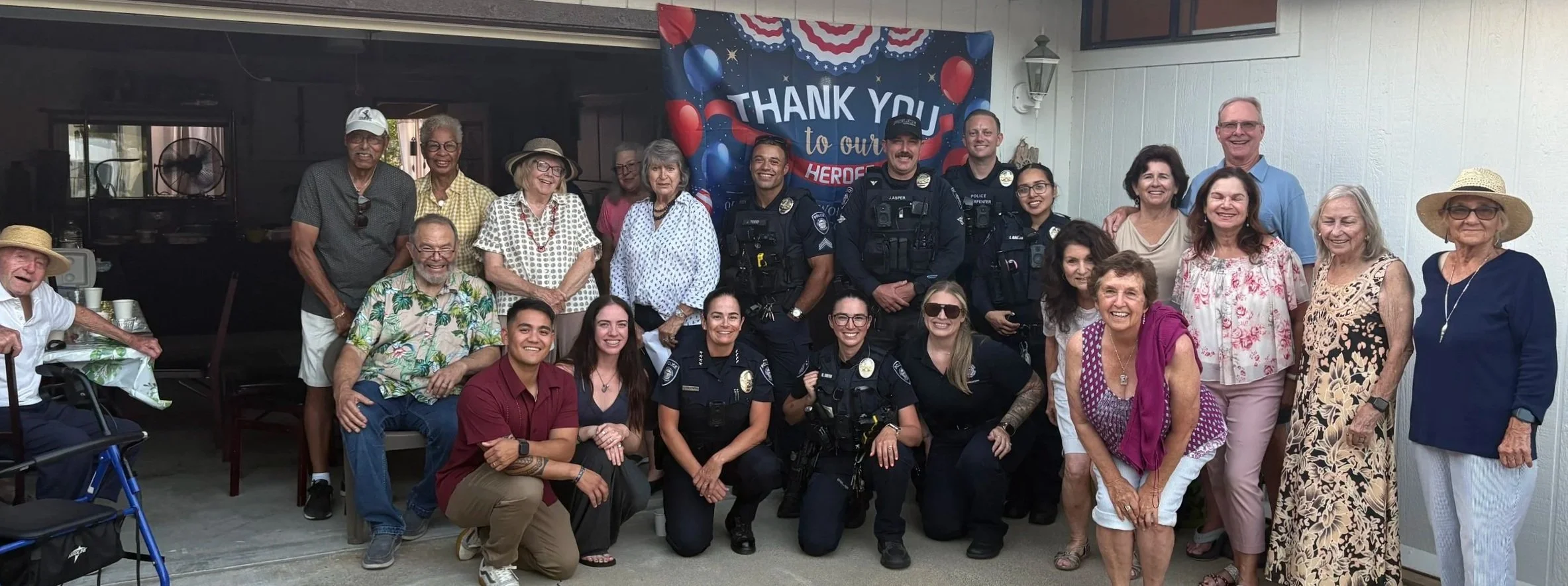 Group of diverse people, including police officers and civilians, gathered outdoors in front of a patriotic 'Thank You' banner with red, white, and blue colors, balloons, and stars, celebrating a community event.