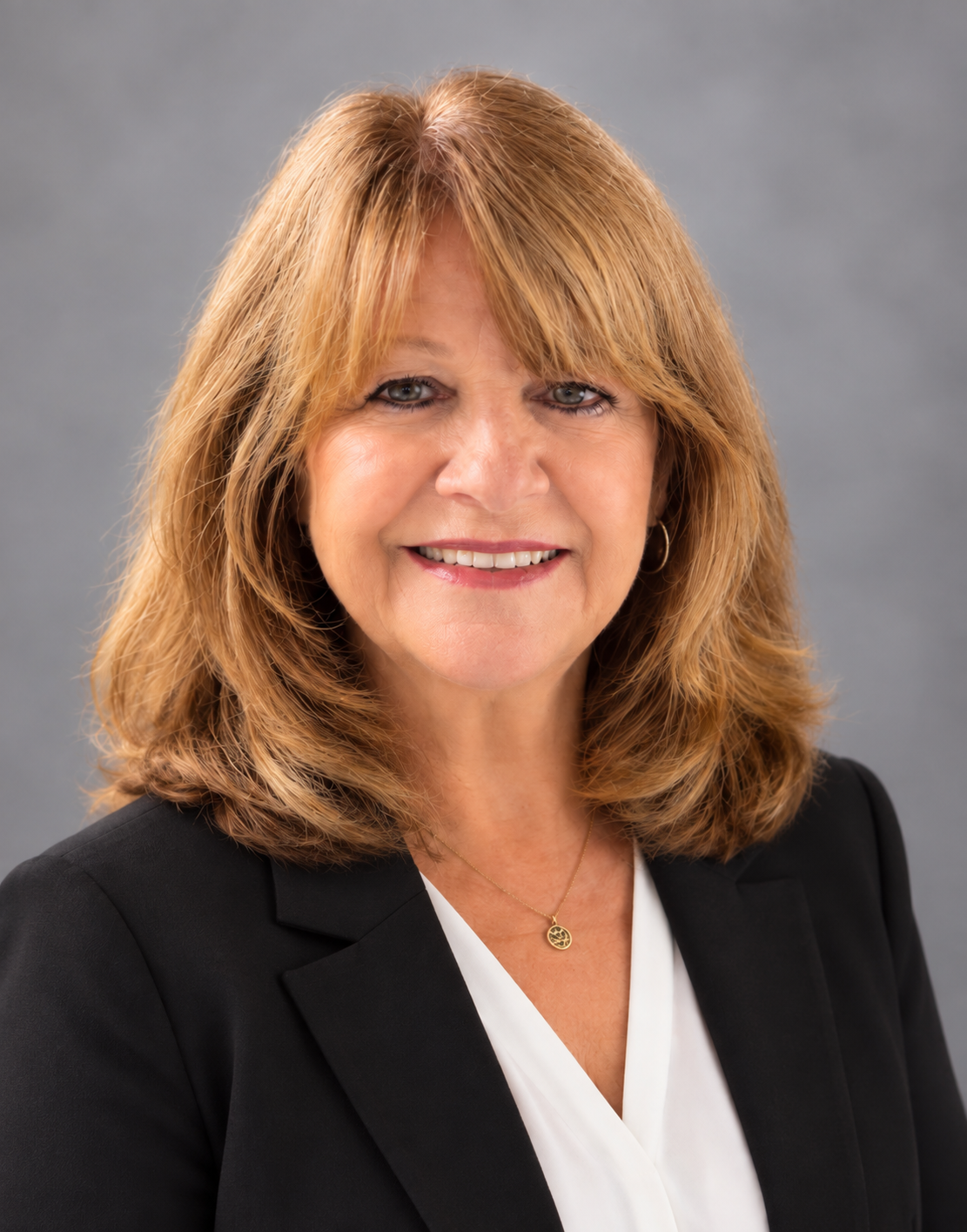 Camino Hills Real Estate Expert. Professional headshot of a smiling middle-aged woman with shoulder-length reddish-blonde hair, wearing a black blazer, white blouse, and a gold necklace, against a gray background.