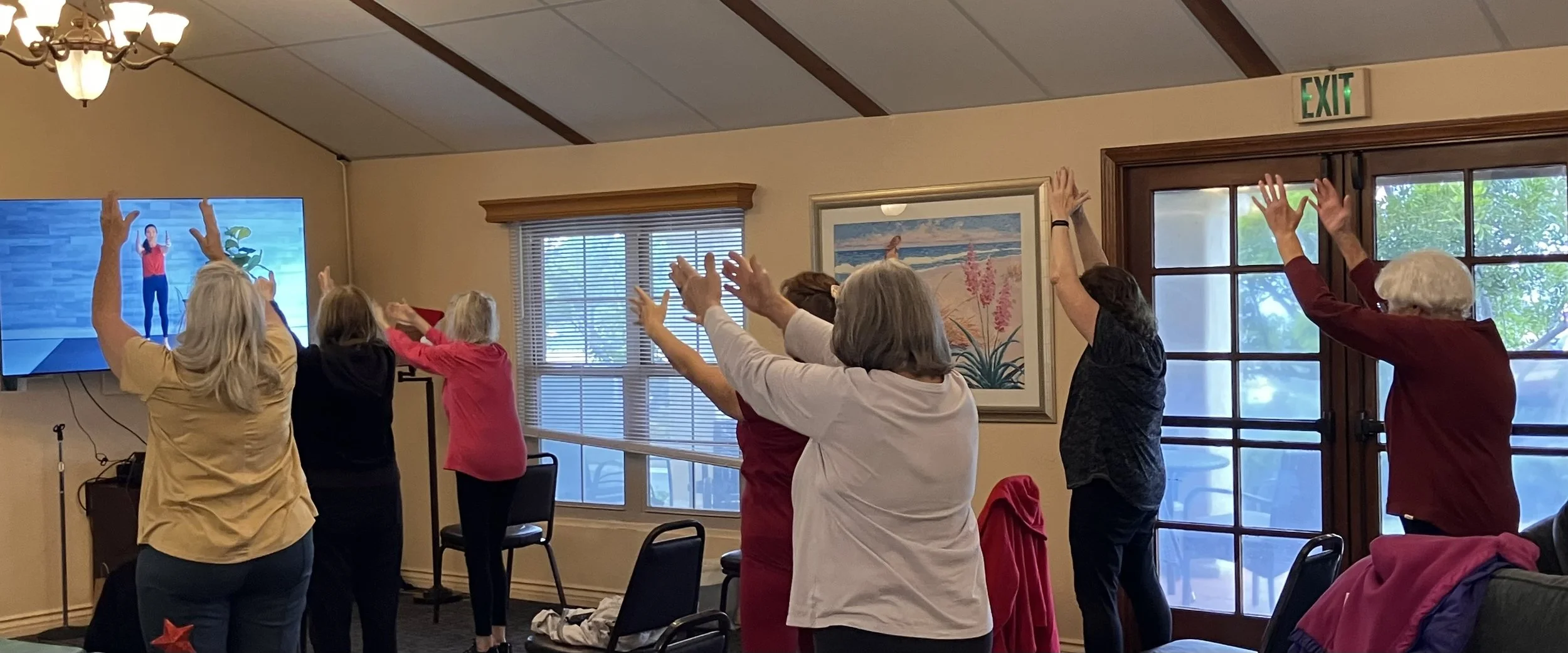 Group of people participating in a seated exercise class led by an instructor on a screen, with participants raising their arms in a warm-up or stretching routine inside a room with windows, artwork, and a door.