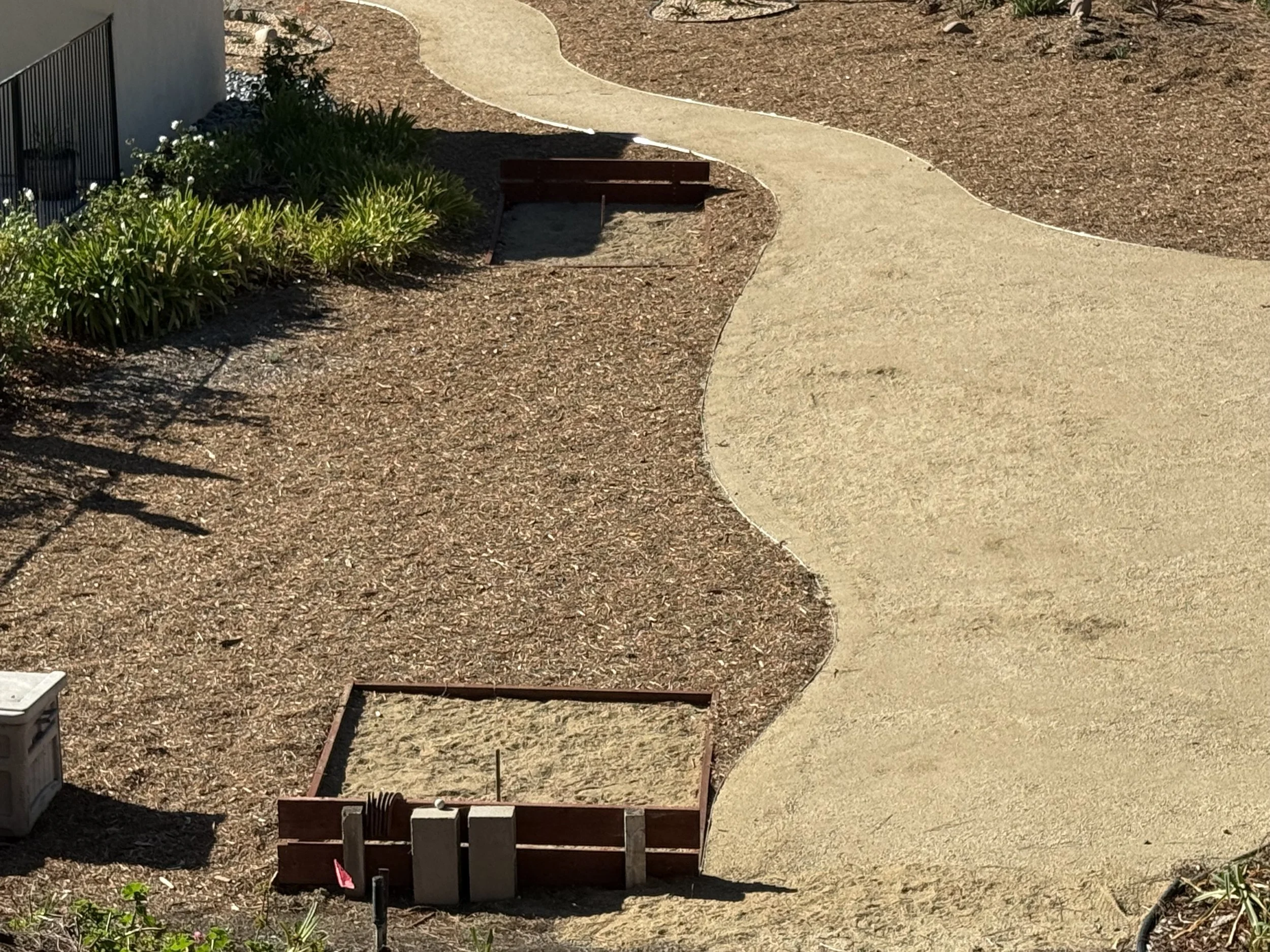 A winding concrete sidewalk cutting through a landscape with dirt and sparse greenery, bordered by garden beds with shrubs, and some construction frames or boxes in the foreground.