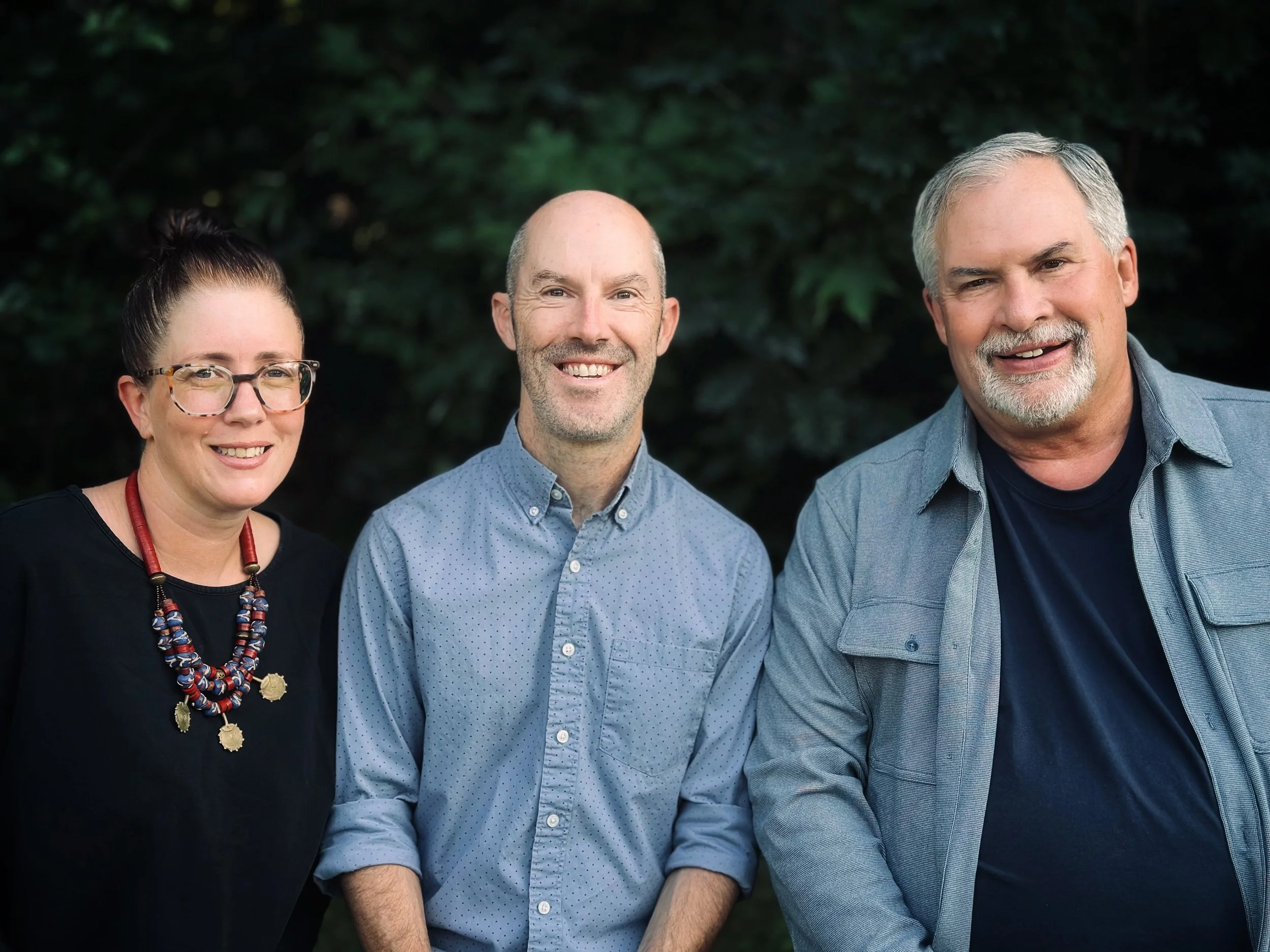 Three adults smiling outdoors against a background of green foliage. The woman on the left has dark hair in a bun, glasses, and is wearing a black top with a colorful beaded necklace. The man in the middle has a shaved head, stubble, and is wearing a light blue, dotted shirt. The man on the right has gray hair, a beard, and is wearing a denim jacket over a dark shirt.