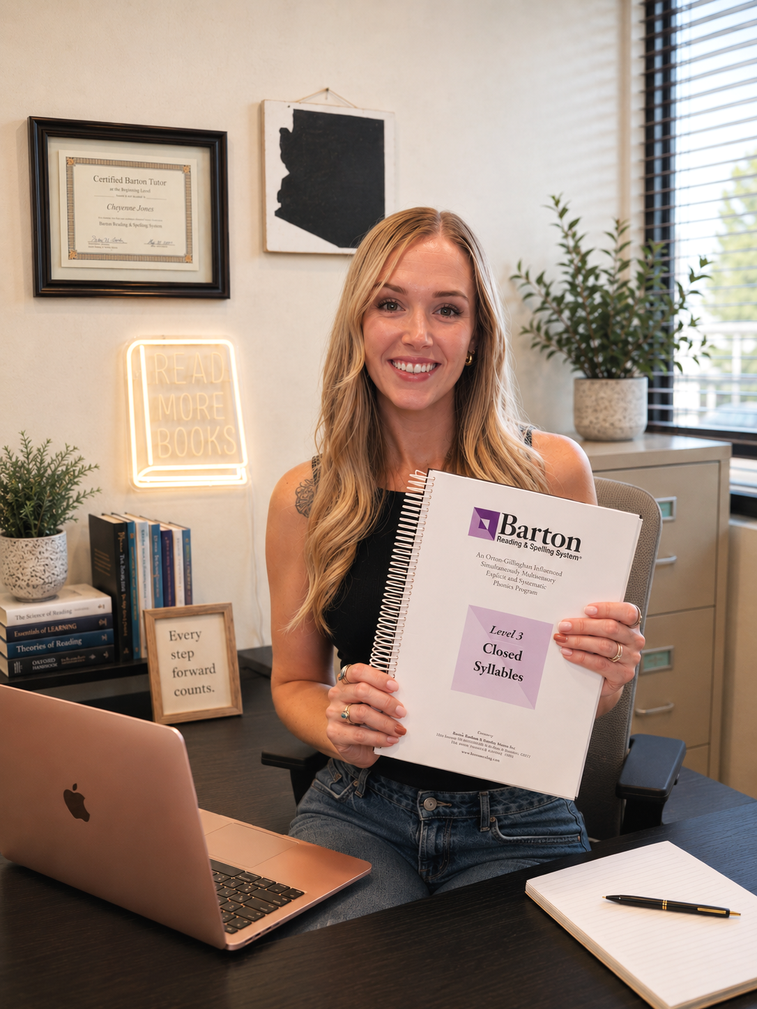 A woman with long blonde hair sitting at a desk in an office, holding a textbook titled 'Barton Reading & Spelling System, Level 3, Closed Syllables,' smiling at the camera.