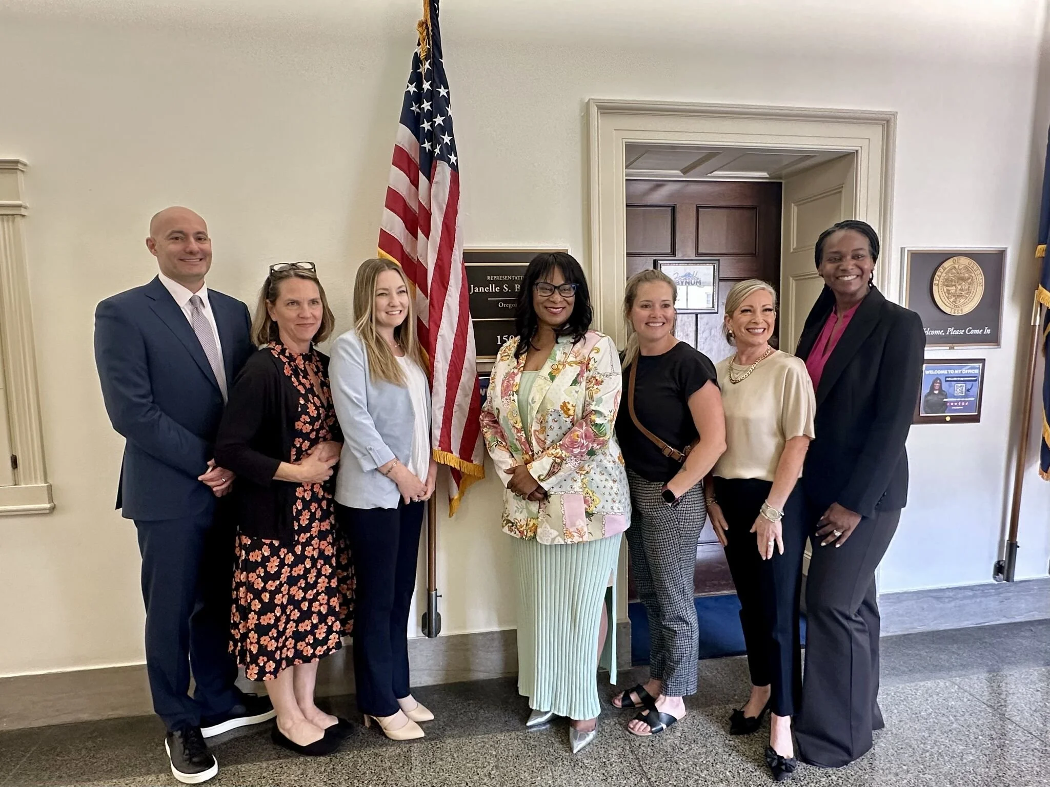 A group of seven diverse people standing together and smiling in front of a sign and an American flag. They are dressed in professional attire, standing inside a building with cream-colored walls and wood accents.