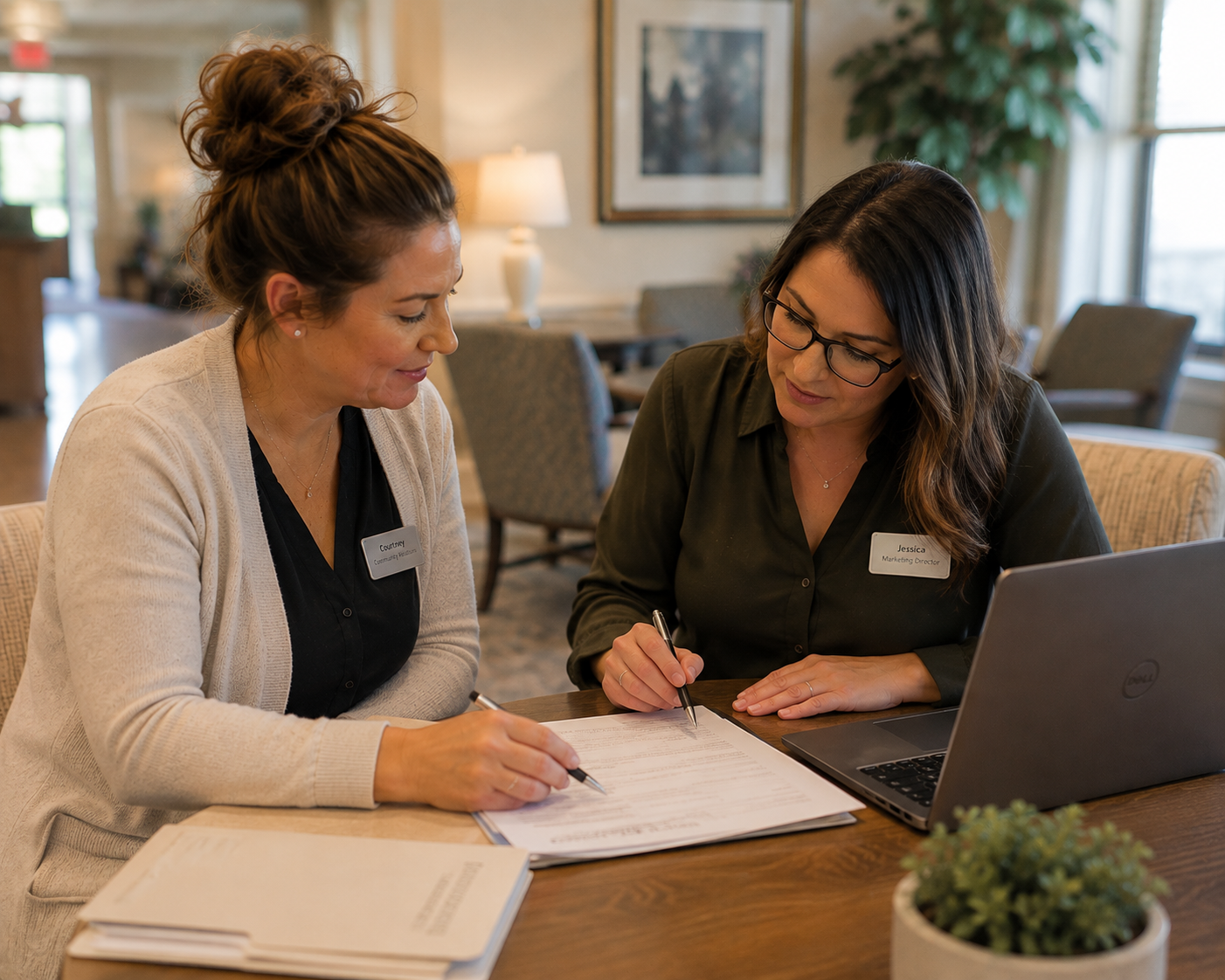 Two women sitting at a table signing documents while looking at each other, with a laptop, books, and a plant in the foreground in a cozy room.