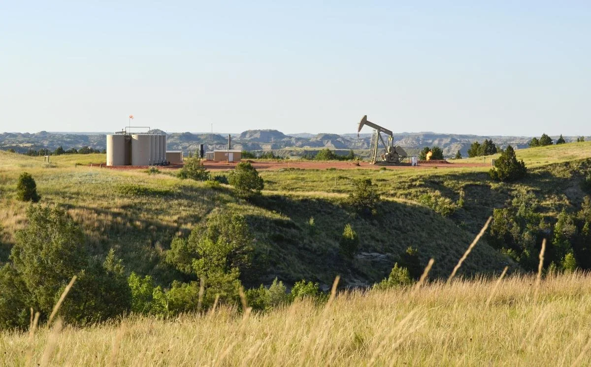 Oil pump jack and storage tanks on a grassy plain with hills in the background under clear sky.