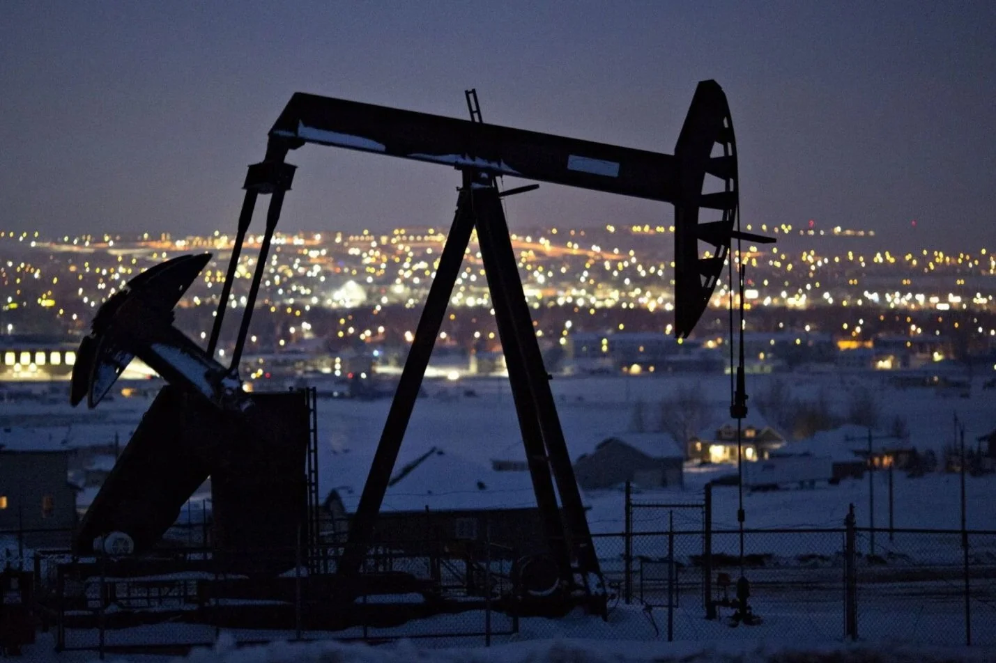 Silhouette of oil pumpjack at night with city lights in the background.