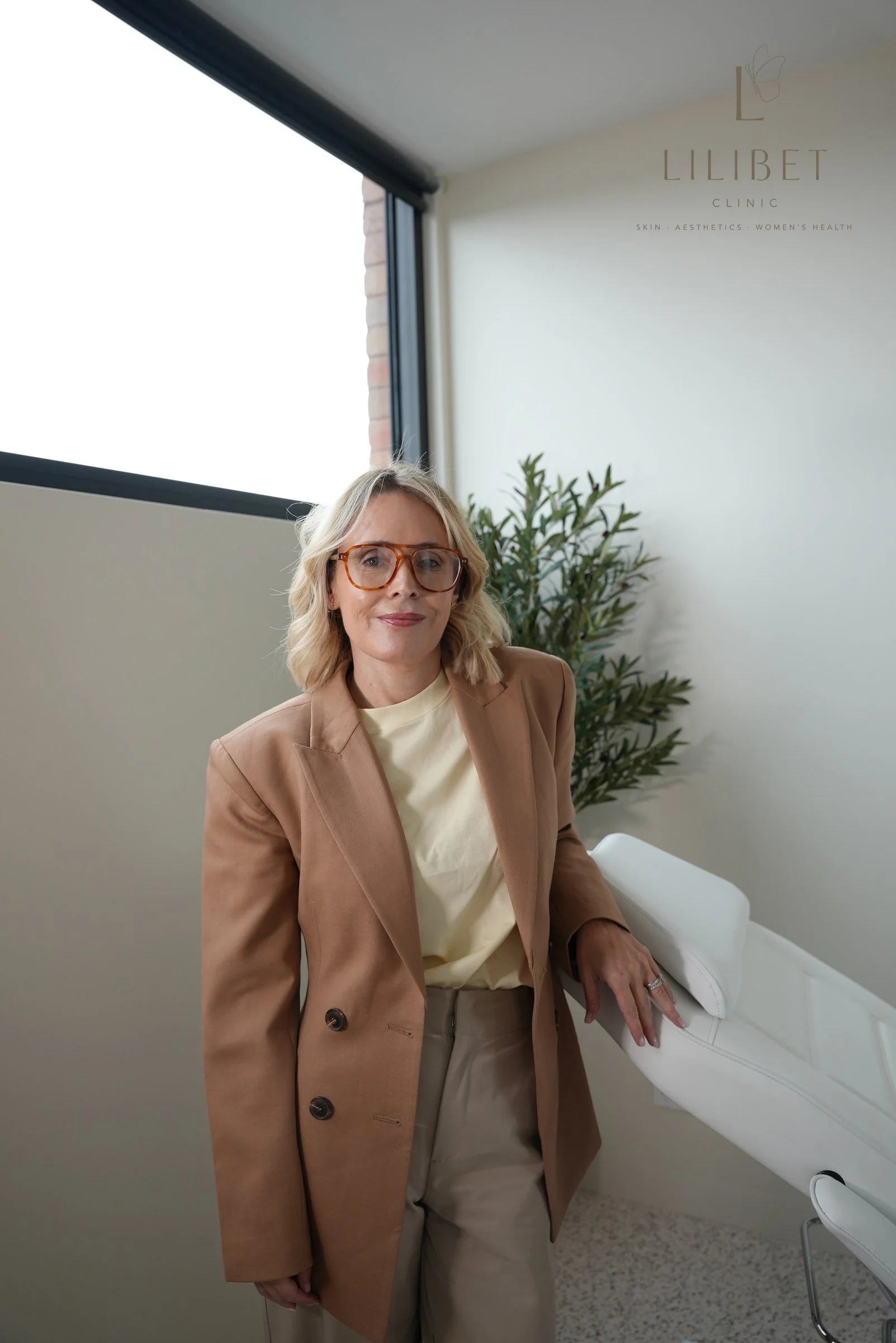 A woman with blonde hair and glasses, wearing a tan blazer and beige pants, standing in a medical or skincare clinic, placing her hand on a white examination bed. The clinic's logo and name, 'Lilibet Clinic,' along with the words 'Skin, Aesthetics, Women's Health,' are visible on the wall behind her, near a window with daylight coming through.