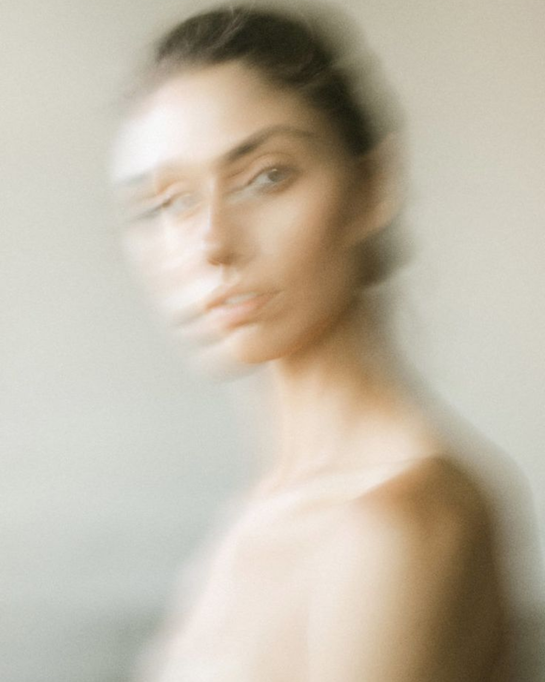 A blurry portrait of a woman with short dark hair, looking to the side, against a plain background.