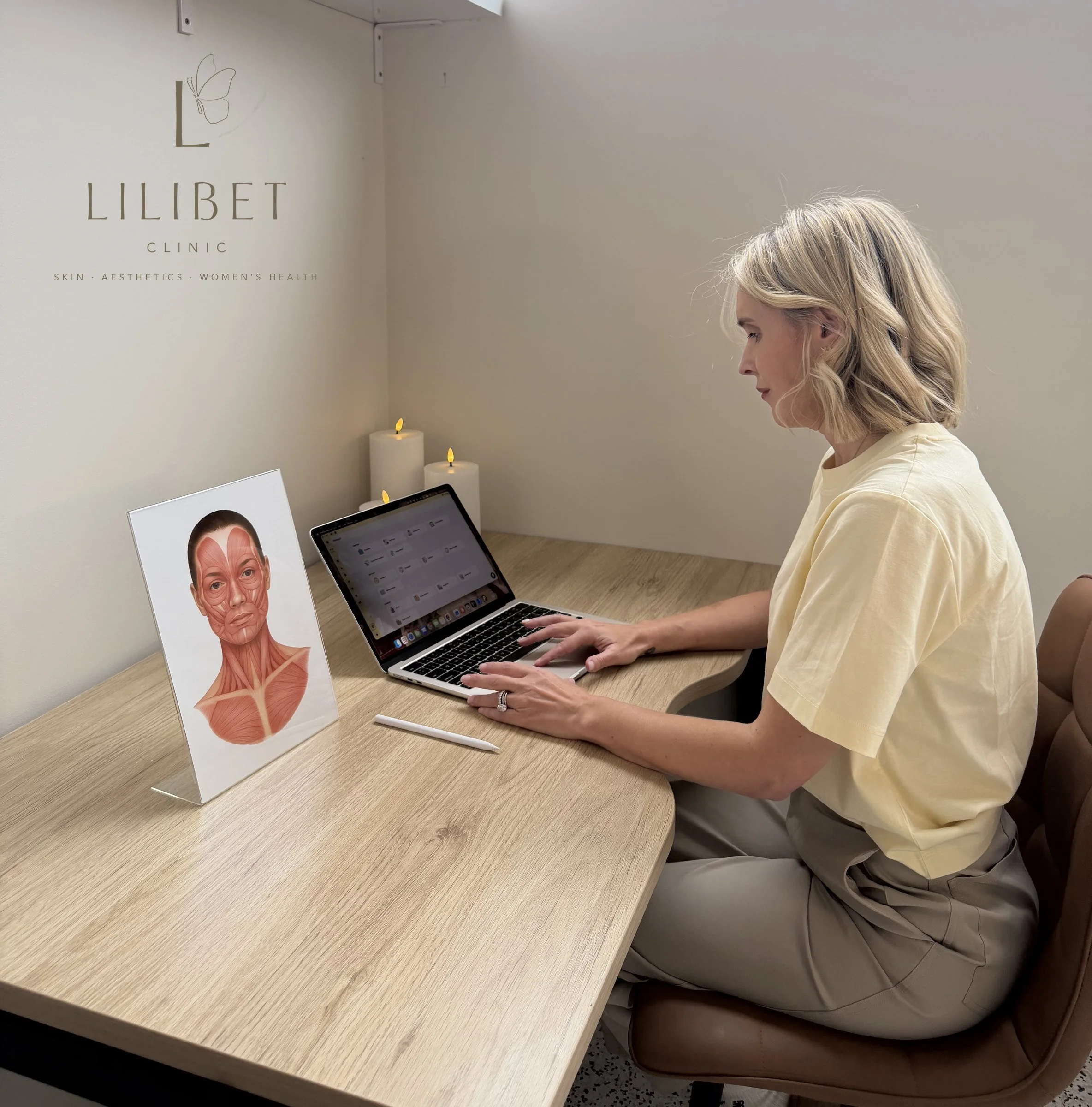 A woman with blonde hair sitting at a wooden table in a skin care clinic. There is a mental health and aesthetics sign on the wall behind her, and a computer in front of her. The setting appears to be a consultation or treatment room.