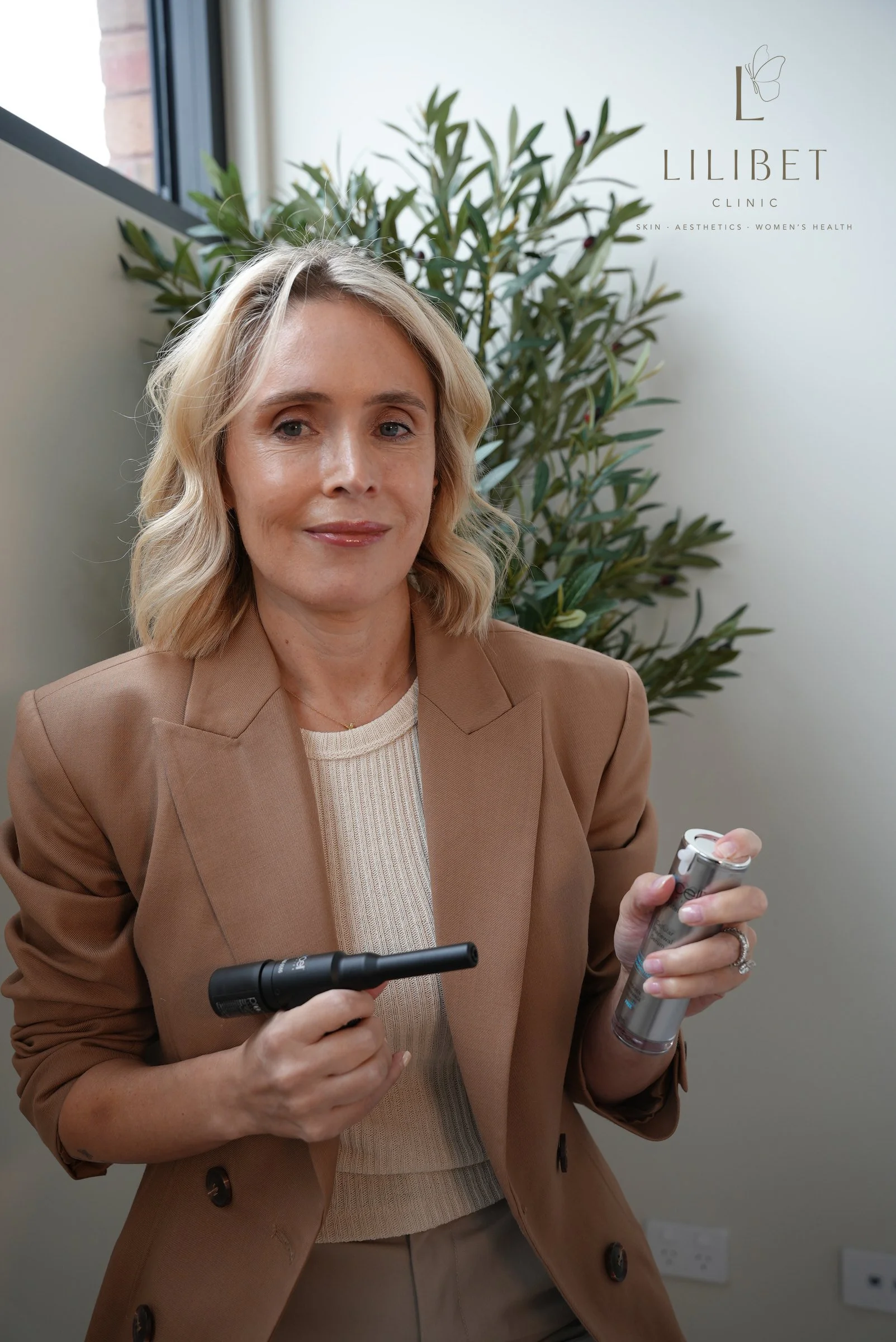 A woman in a tan blazer holding a device in her right hand and a tube in her left hand, standing indoors near a window with a large green plant in the background, and a sign with the text 'LILIBET CLINIC' and a butterfly logo.