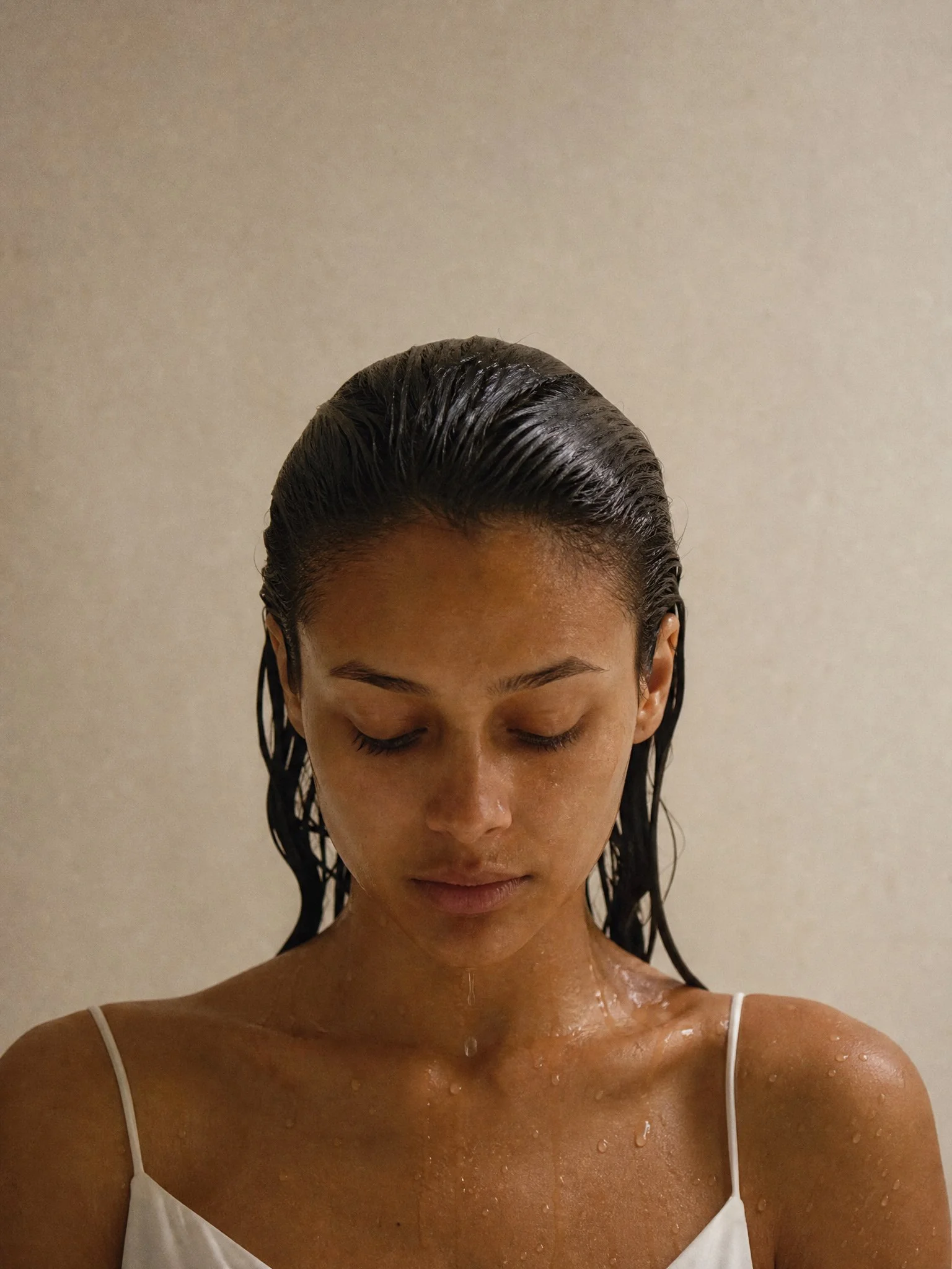 A woman with wet, slicked-back hair and water droplets on her shoulders and face, wearing a white spaghetti strap top, with eyes closed.