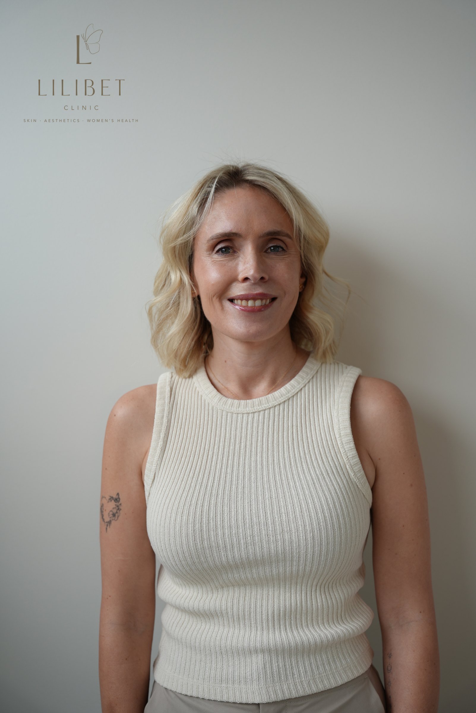 A smiling woman with blonde hair and light skin is standing against a plain wall with the logo of LiliBET Clinic, which specializes in skin, aesthetics, and women's health, visible in the background.