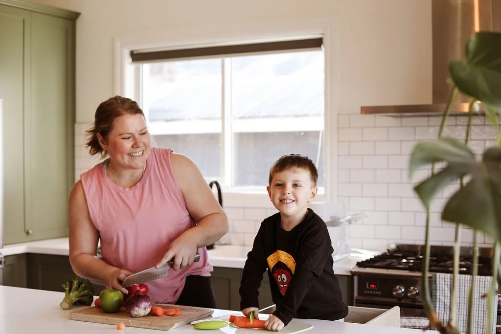 A woman and a young boy in a kitchen preparing vegetables, with the woman smiling and the boy holding a carrot.
