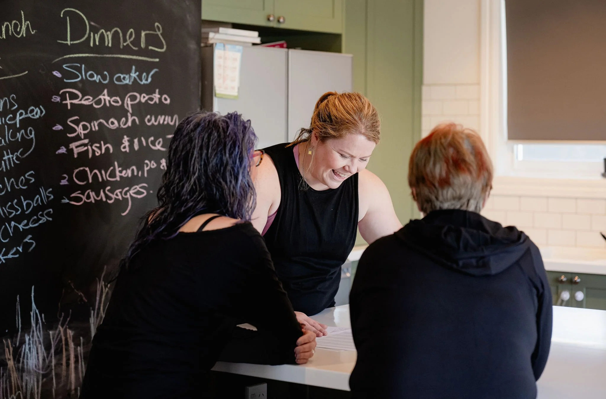 A group of women standing at a kitchen counter, smiling and engaging in conversation, with a chalkboard menu of dinner options in the background.