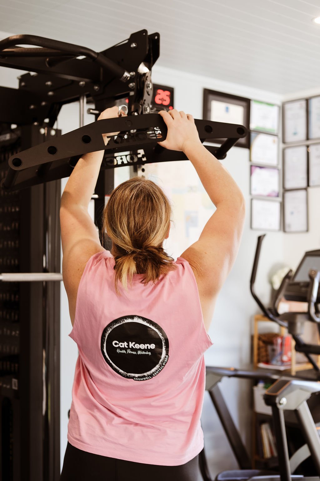 A woman with light brown hair tied in a small braid, wearing a pink sleeveless shirt with a 'Cat Keene' logo on the back, is working out by pulling on a black pull-up bar in a gym. The gym has framed pictures on the wall and exercise equipment.