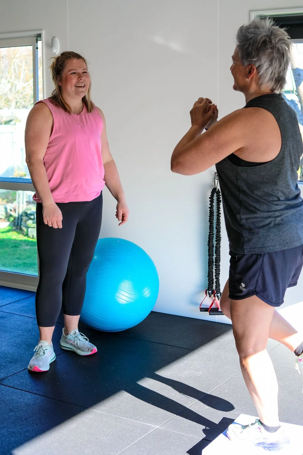 A woman in pink athletic wear laughing while a man in dark clothing exercises nearby in a bright home gym with large windows, a blue exercise ball, and workout equipment.