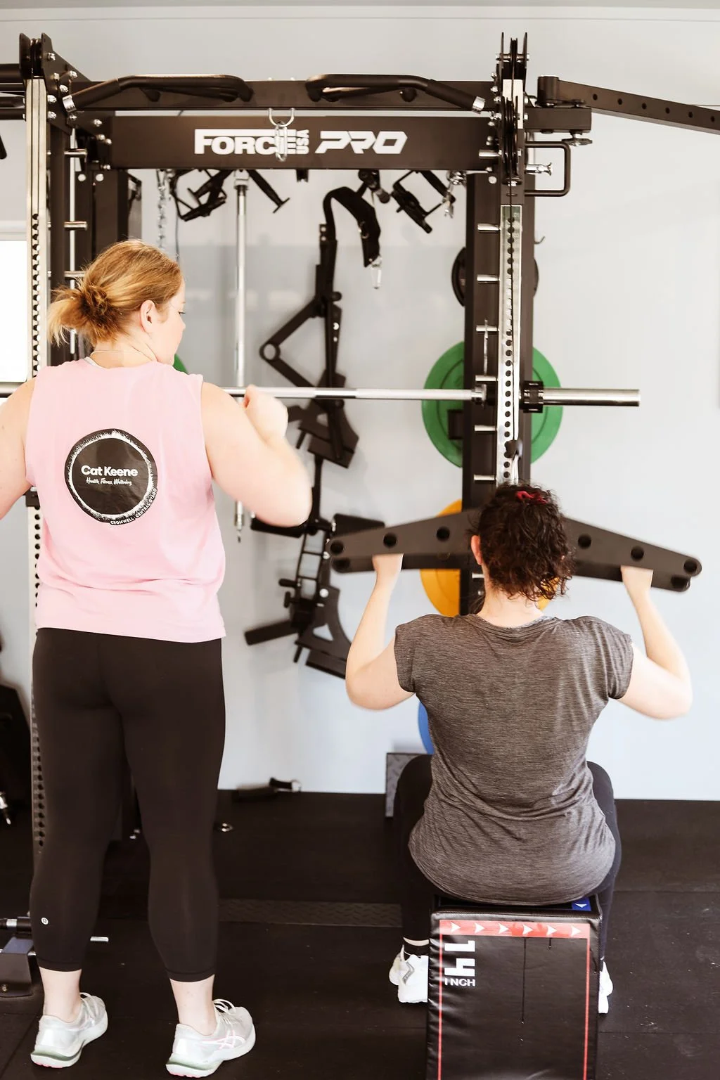 A woman assisting a person with curly hair in lifting a barbell at a gym.