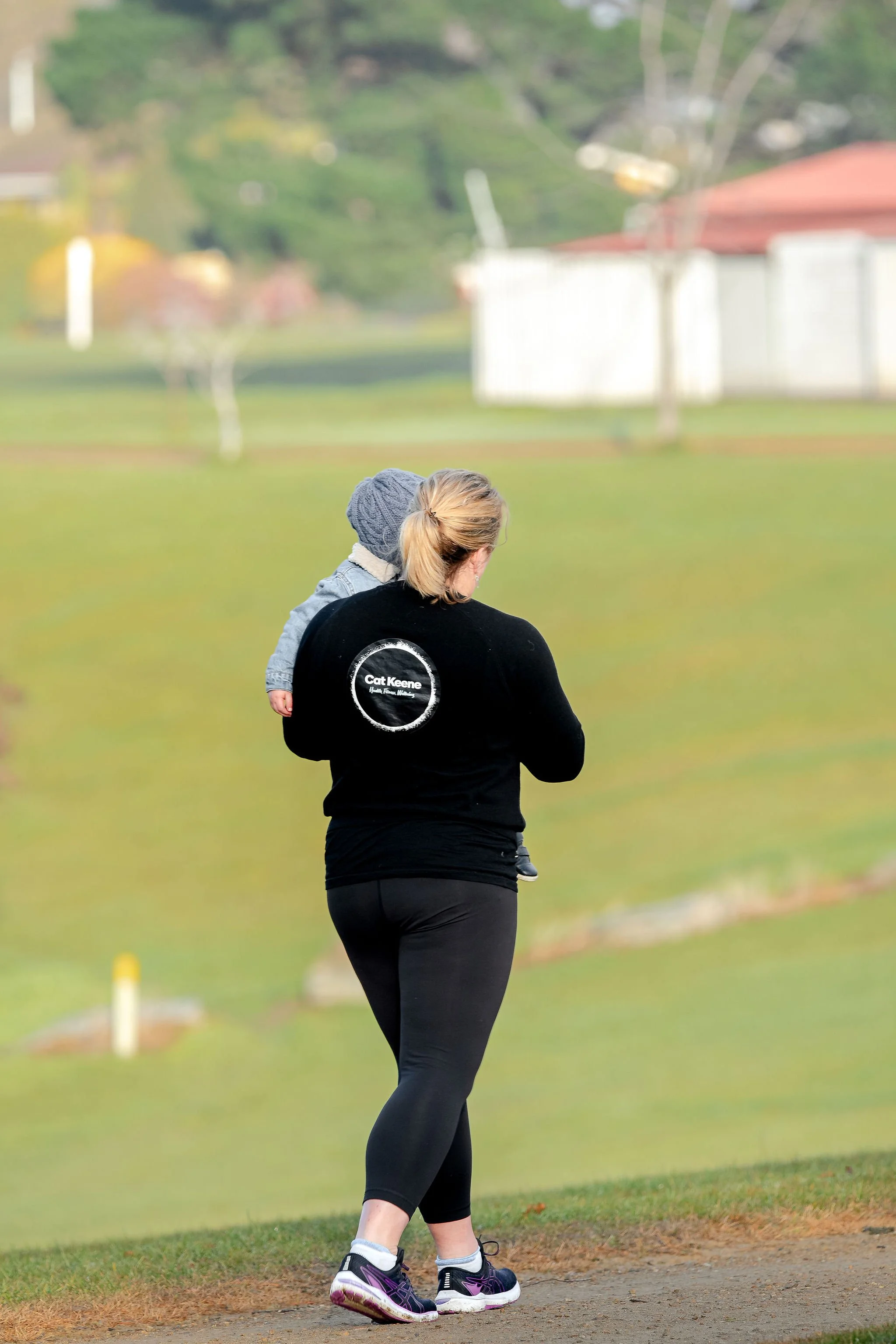 A woman with blonde hair tied in a ponytail, wearing black athletic leggings, a black jacket with a logo on the back, and purple running shoes, is walking in a park with a small child in her arms. The child is wearing a gray knit hat, a gray coat, and dark pants. The background features a grassy area, trees, and a white fence, with a slightly blurred scene.