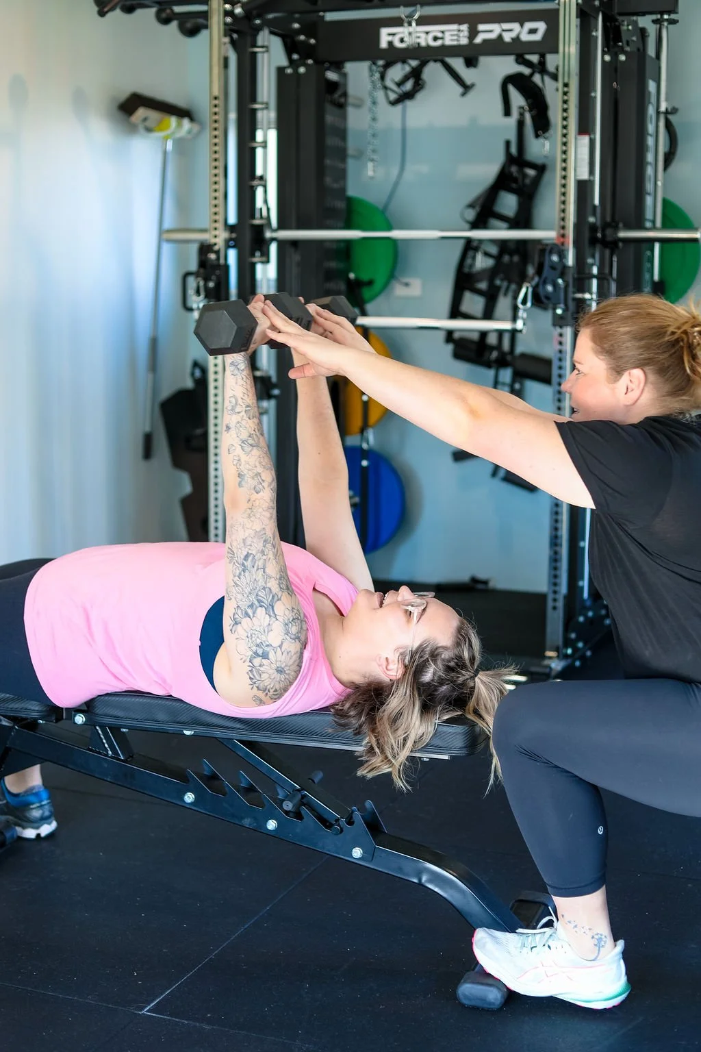 Woman lying on a workout bench, lifting a dumbbell, assisted by a trainer in a gym.