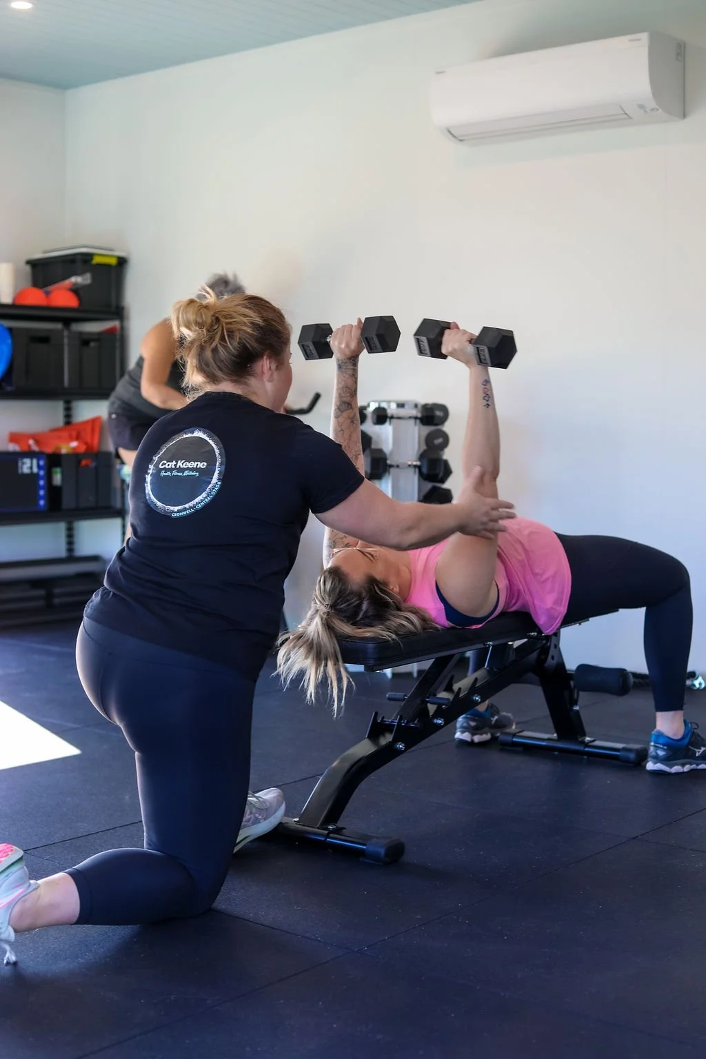 A woman is lying on a workout bench lifting dumbbells with assistance from a trainer kneeling beside her in a gym.