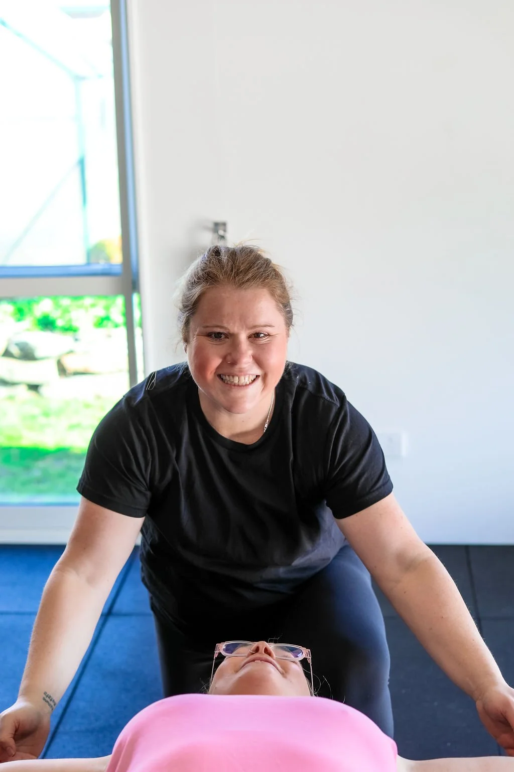 A woman smiling while leaning over a person lying on a table, possibly during a massage or therapy session, in a room with a sliding glass door showing greenery outside.
