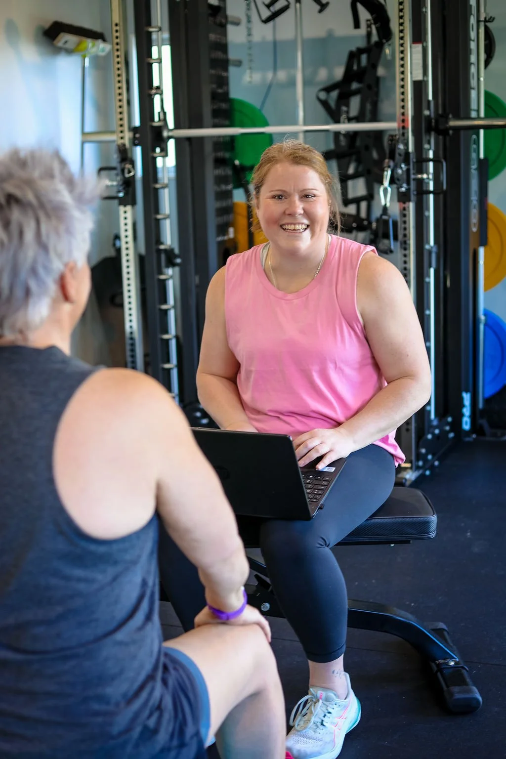 A woman in a pink tank top smiling while talking to a man with white hair wearing a black tank top at a gym, with workout equipment in the background.