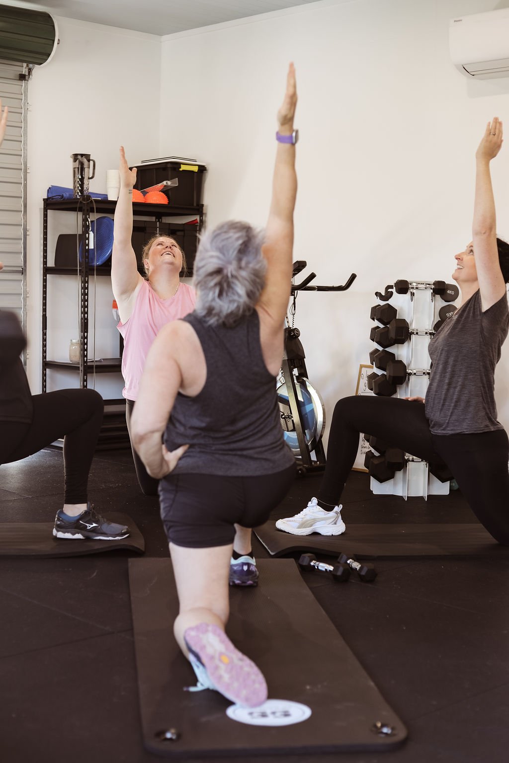 Three women performing a yoga pose together in a gym, with one woman kneeling and the other two standing with one arm raised.