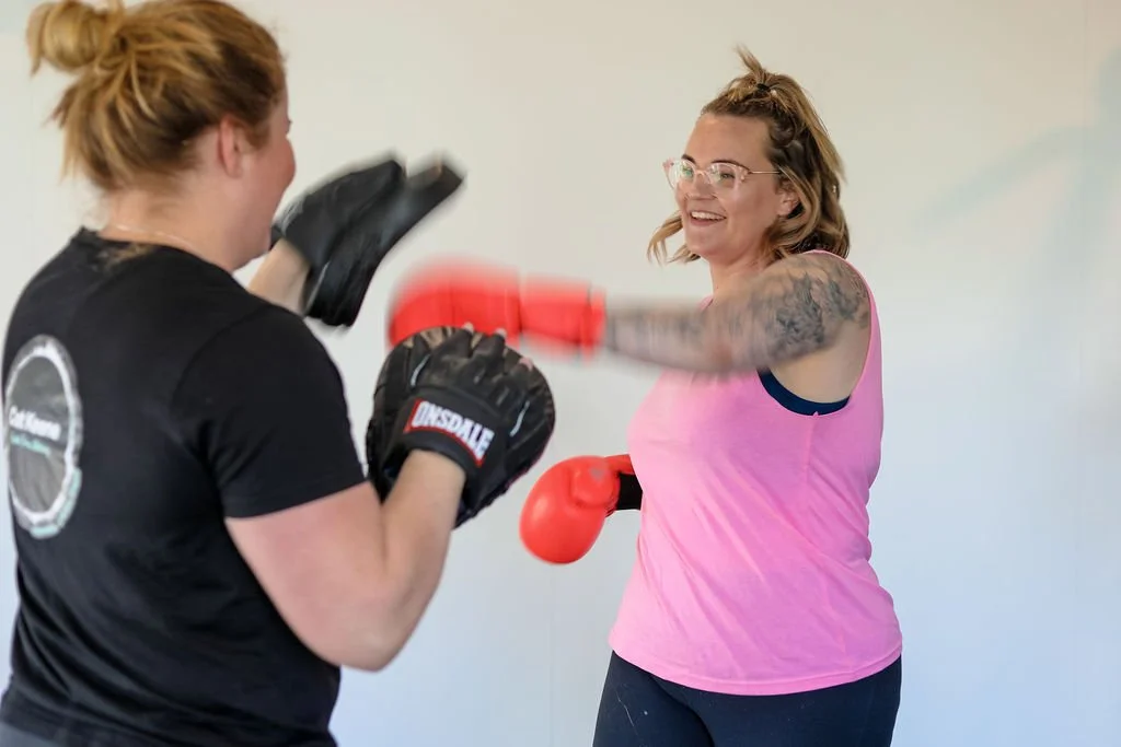 Two women sparring with boxing gloves, one is smiling and wearing a pink tank top, the other has her back to the camera and is wearing a black shirt.