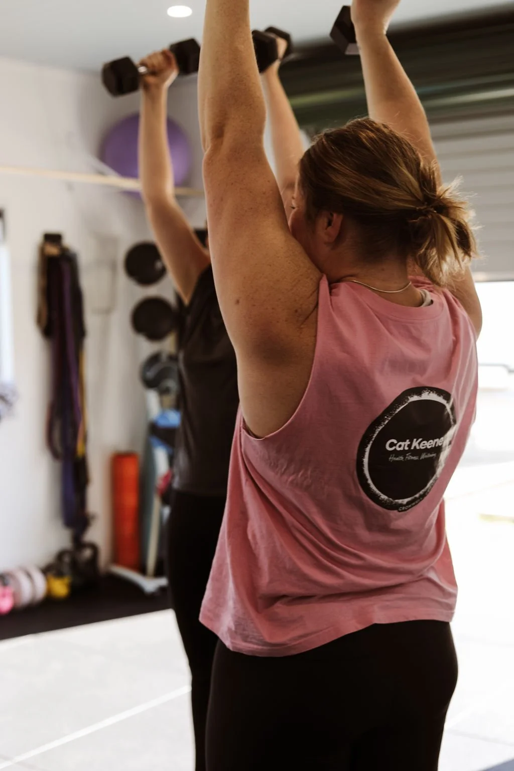 A woman in a pink sleeveless shirt lifting dumbbells in a gym, with another person partially visible in the background.