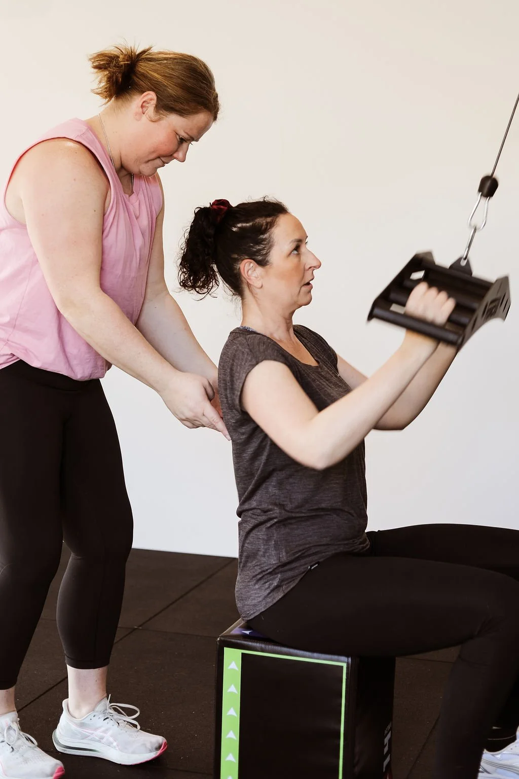 A woman sitting on a workout box using a pulley system to lift weight with her arm, guided by a trainer standing behind her, in a fitness or physical therapy setting.