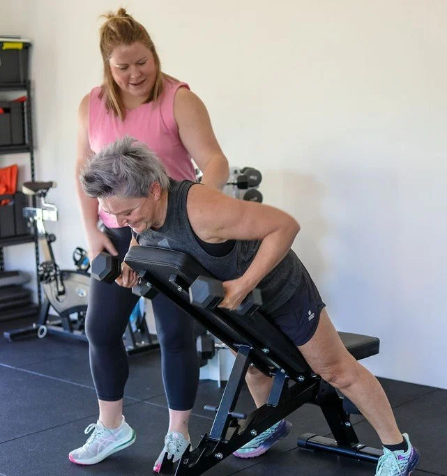 A woman using a dumbbell bench with assistance from a trainer in a gym.