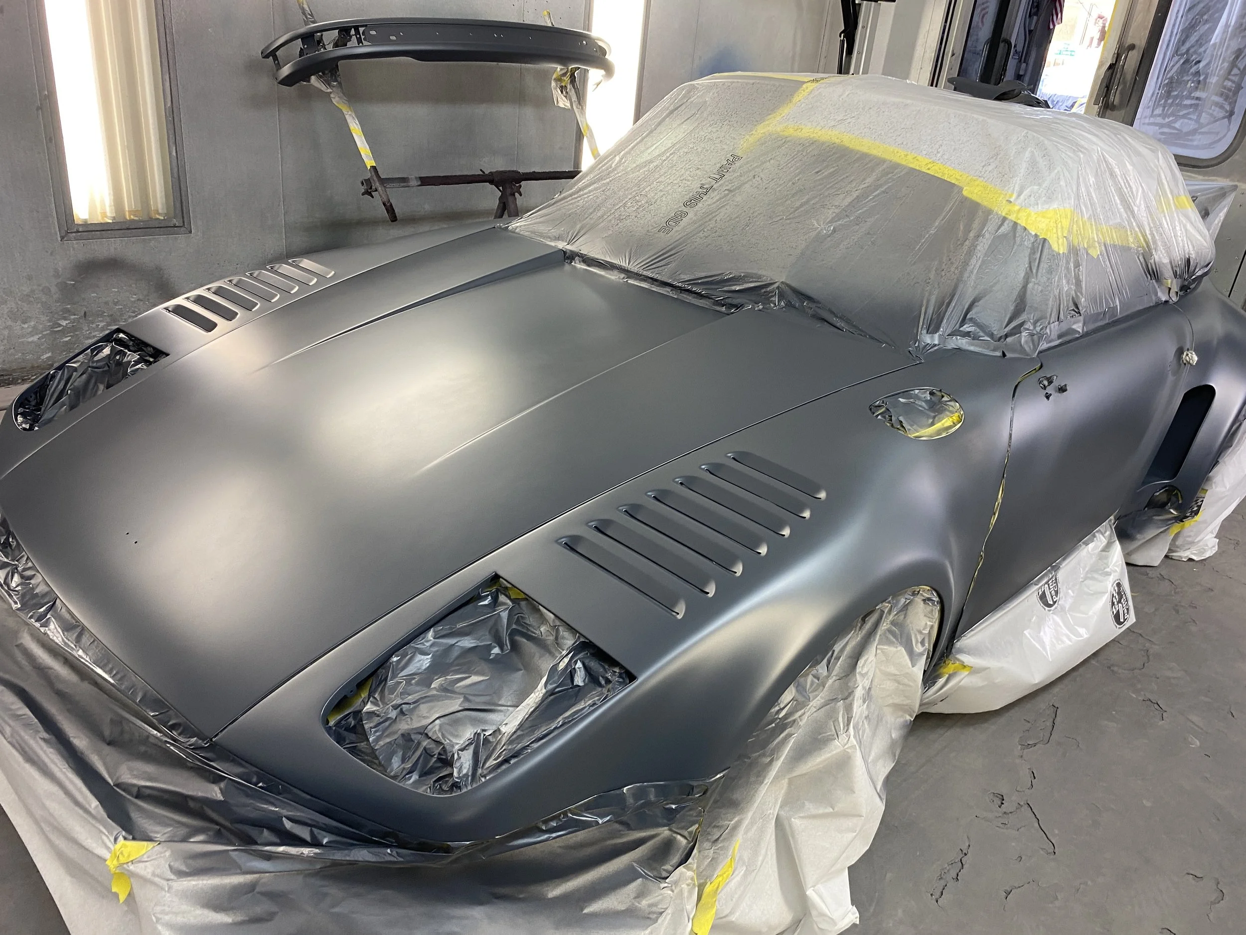 A silver vintage car being painted or repaired in a workshop, covered with protective plastic sheeting and masking tape, with some parts removed and a workspace in the background.