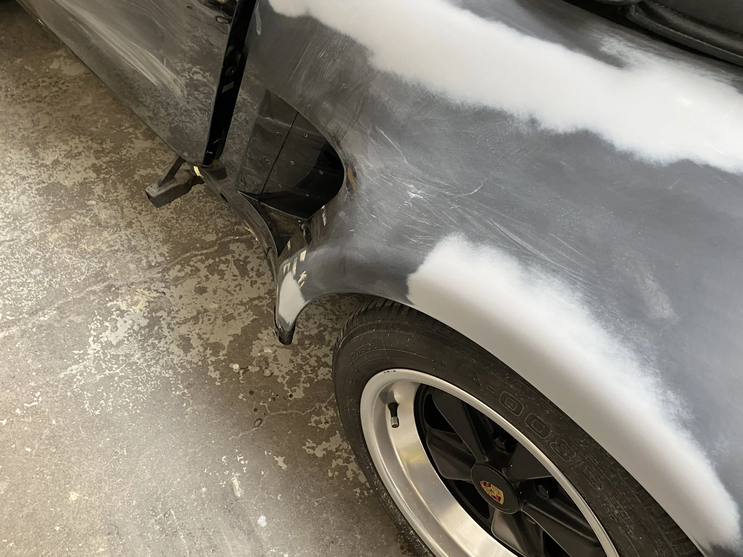 Close-up of a black car's fender with a freshly applied coat of primer, showing some scratches and a wheel with a silver rim and black tire.