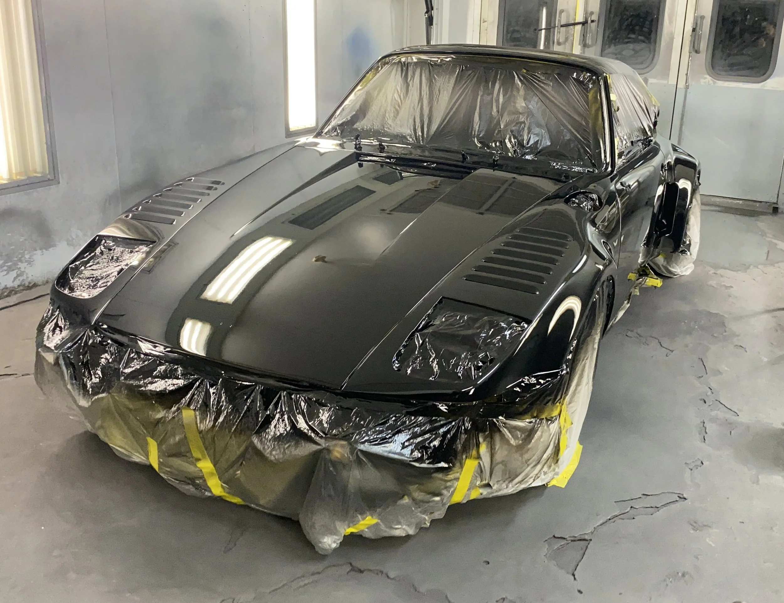 A black sports car covered with protective tape and masking paper, inside a paint booth or garage, preparing for or undergoing paintwork.