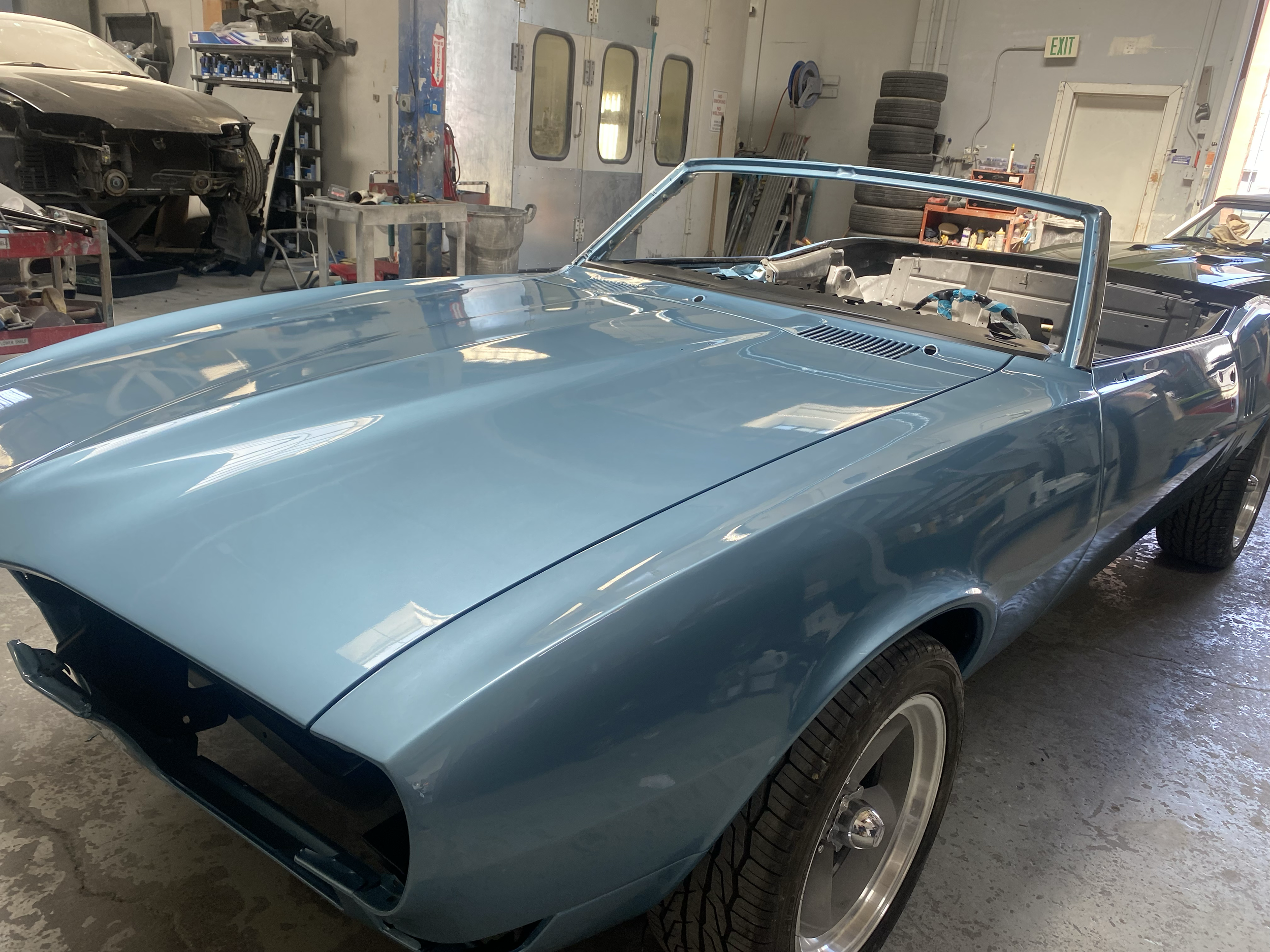 A vintage blue sports car with an open top inside a garage or workshop, surrounded by tools and tires, with a partially disassembled black car in the background.