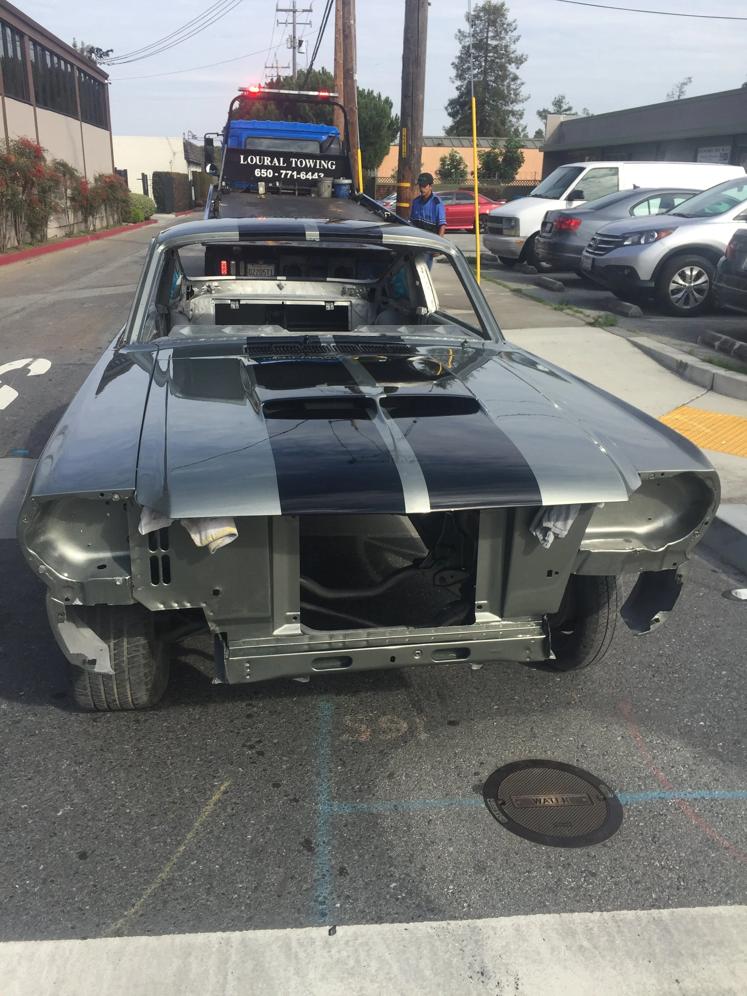 Empty car frame with no doors or interior, parked in a parking lot, with a highway on the side and a tow truck behind it.