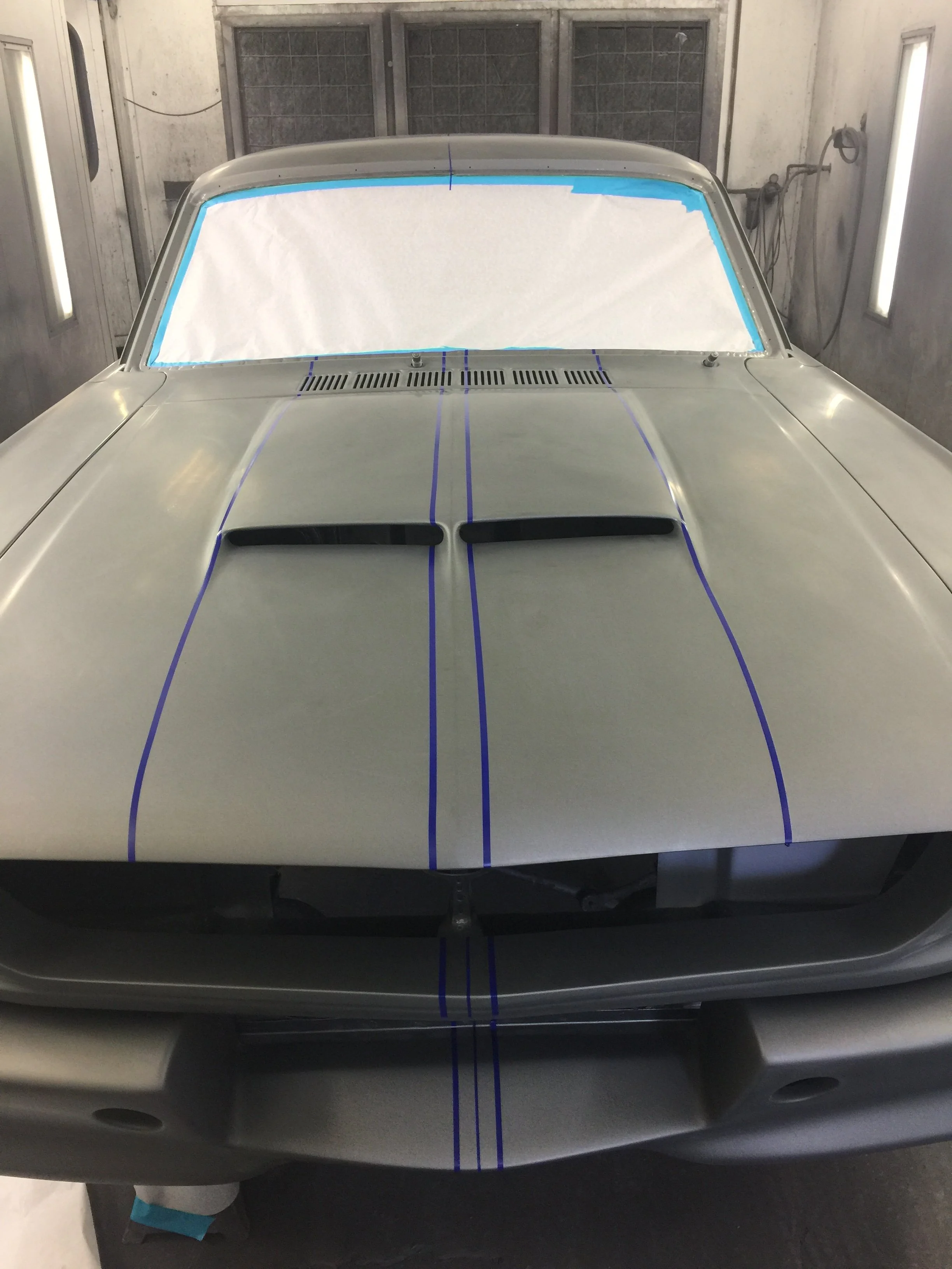 Front view of a silver car in a paint booth with masking tape around the windshield and hood, and masking paper covering the windshield.
