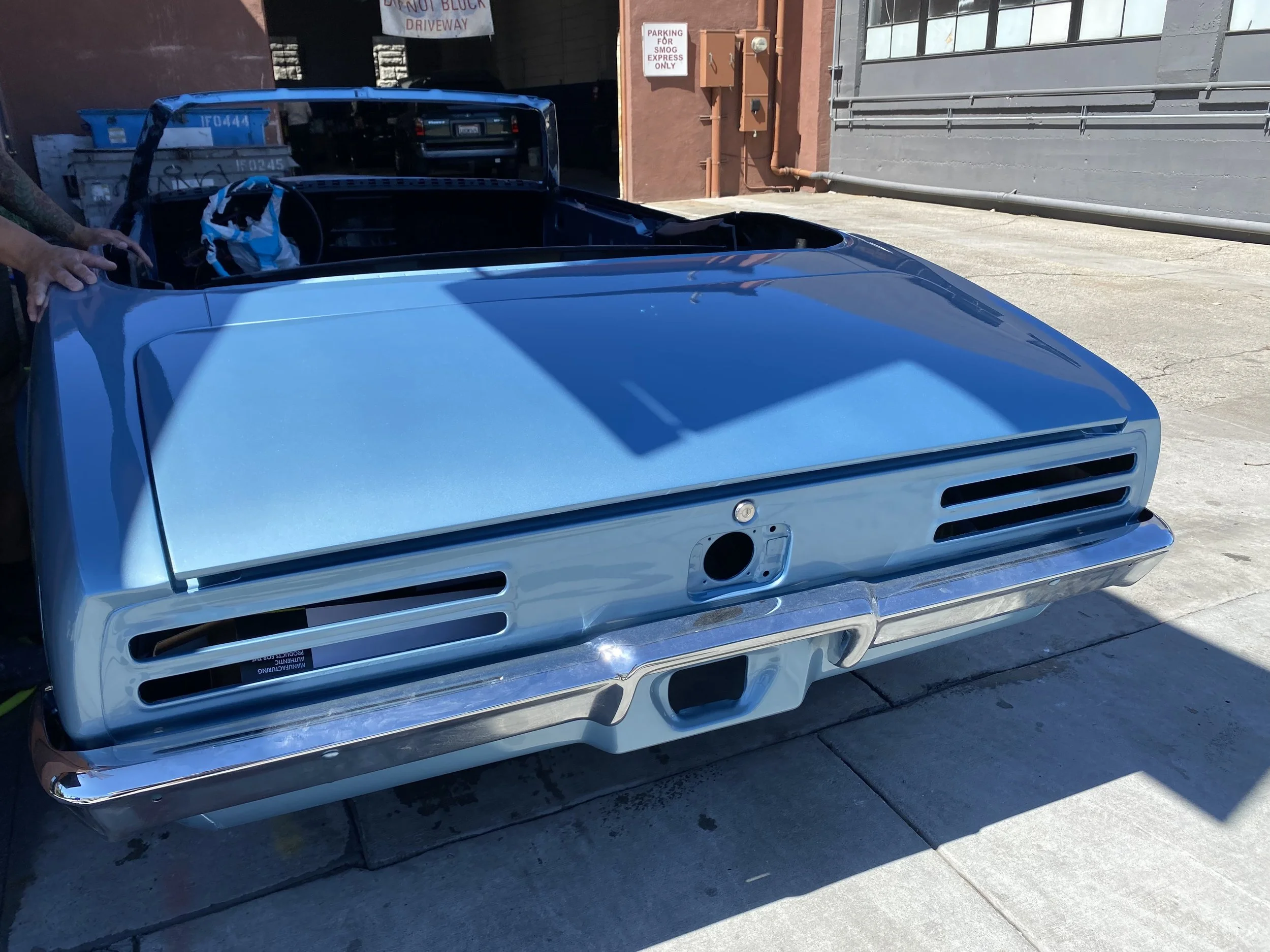 Rear view of a blue vintage car with no license plate, parked outdoors near a building with industrial walls, in front of trash bins, and with a person holding the side of the car visible in the left part of the image.