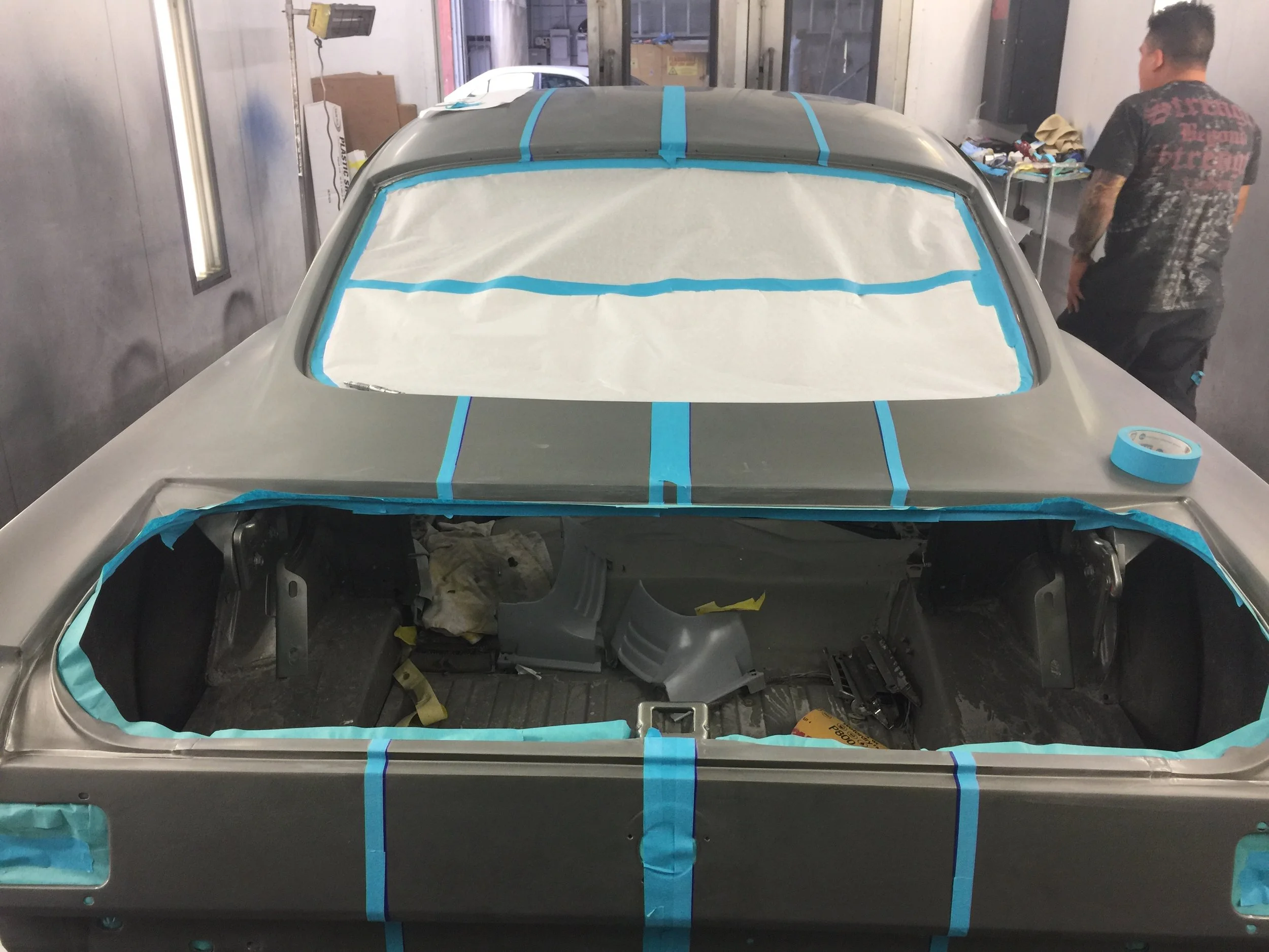 The image shows a car in a paint booth being prepared for painting. The windshield and other parts are covered with masking paper and blue painter's tape to protect areas from paint. A person stands near the car, working on the project.