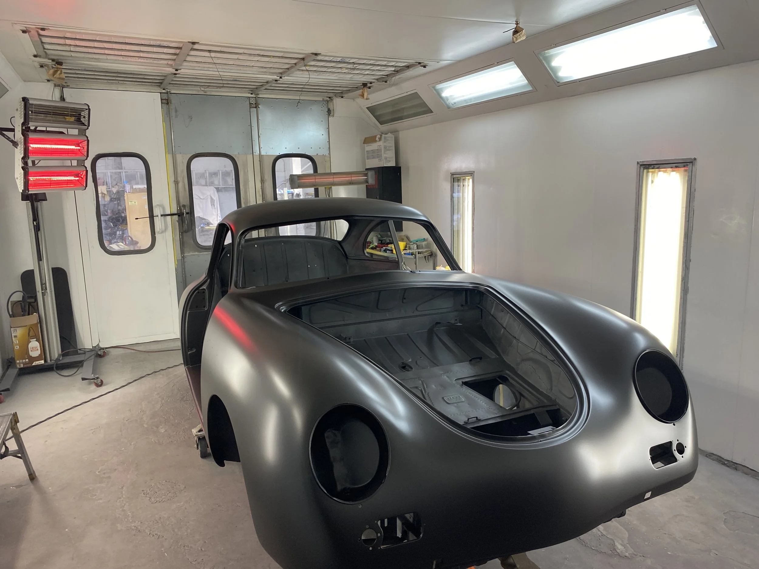 A black car body shell inside a garage or workshop, with no windows or engine, surrounded by tools and equipment, and a ceiling with skylights and fluorescent lights.