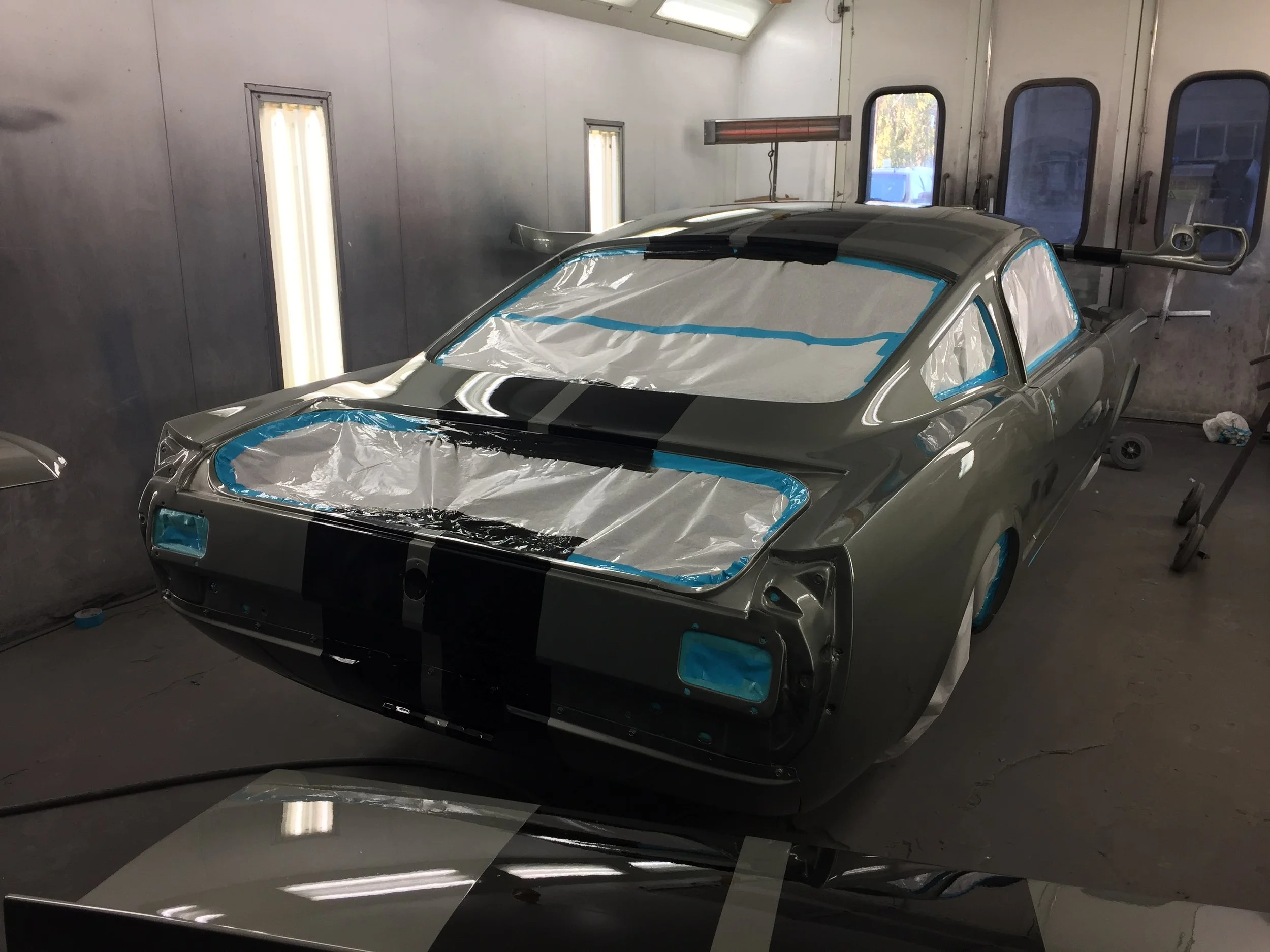 A silver sports car is inside a paint booth, covered with protective foil around the windshield, windows, and part of the body, as it undergoes painting or finishing work.