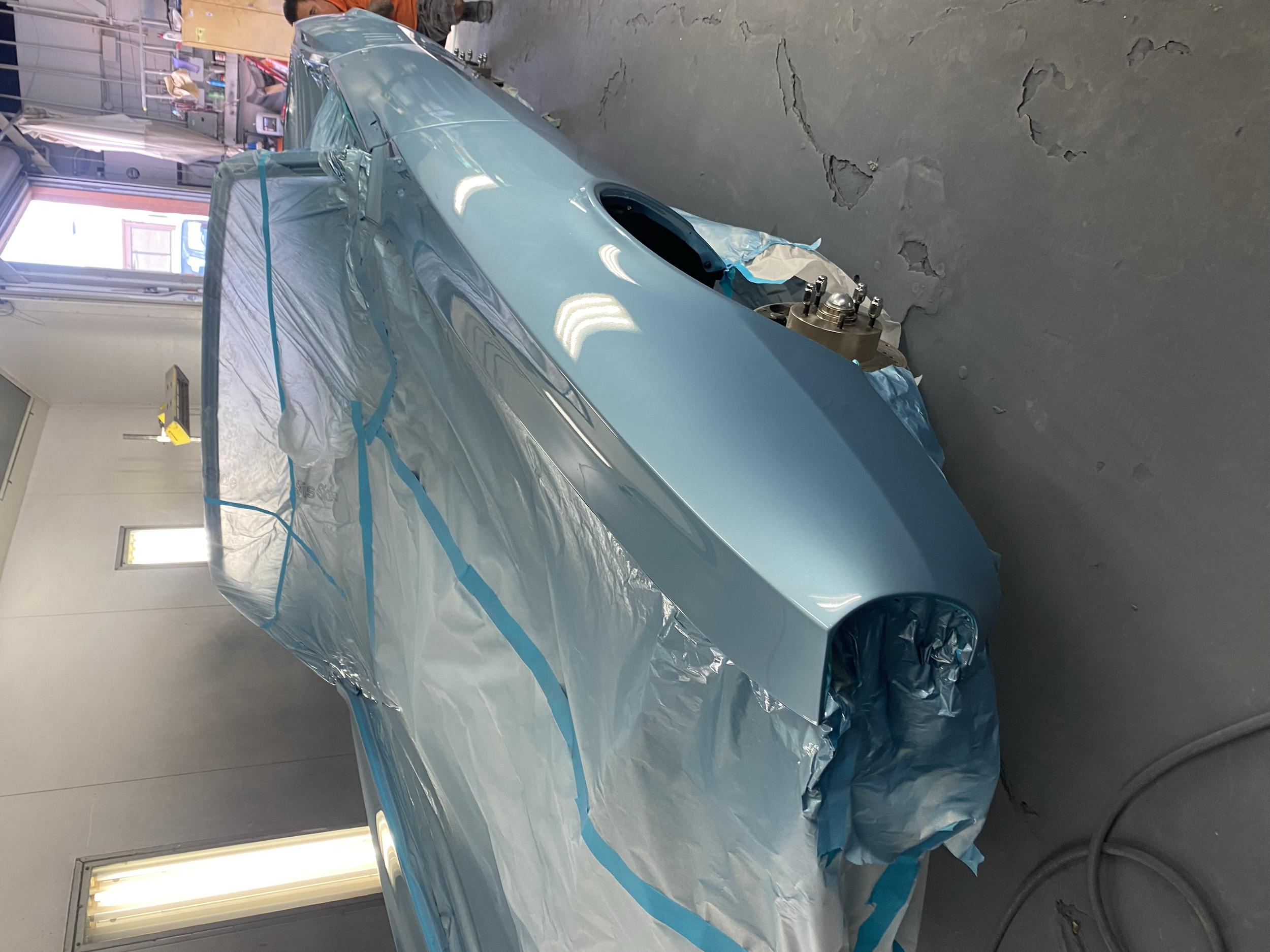 A vintage car is being painted in a workshop, partially covered with blue painter's tape and protective plastic sheeting, with the front wheel removed.