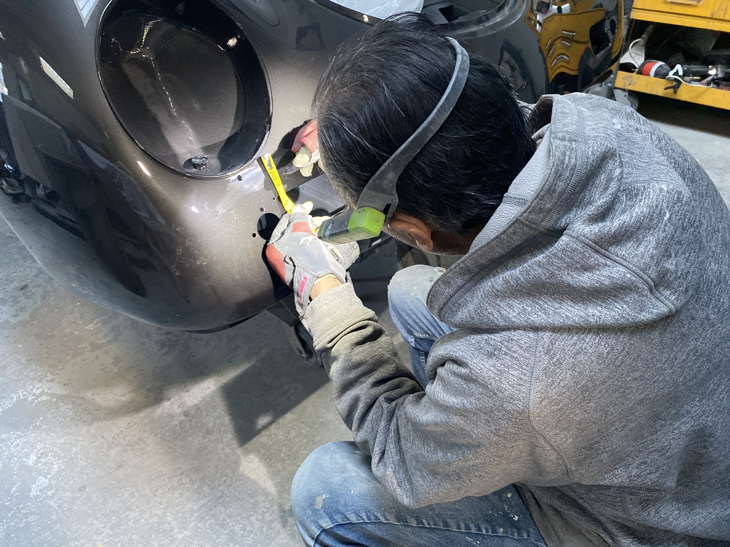 A mechanic working on a black car, wearing gloves and safety glasses, in a garage with tools and equipment nearby.