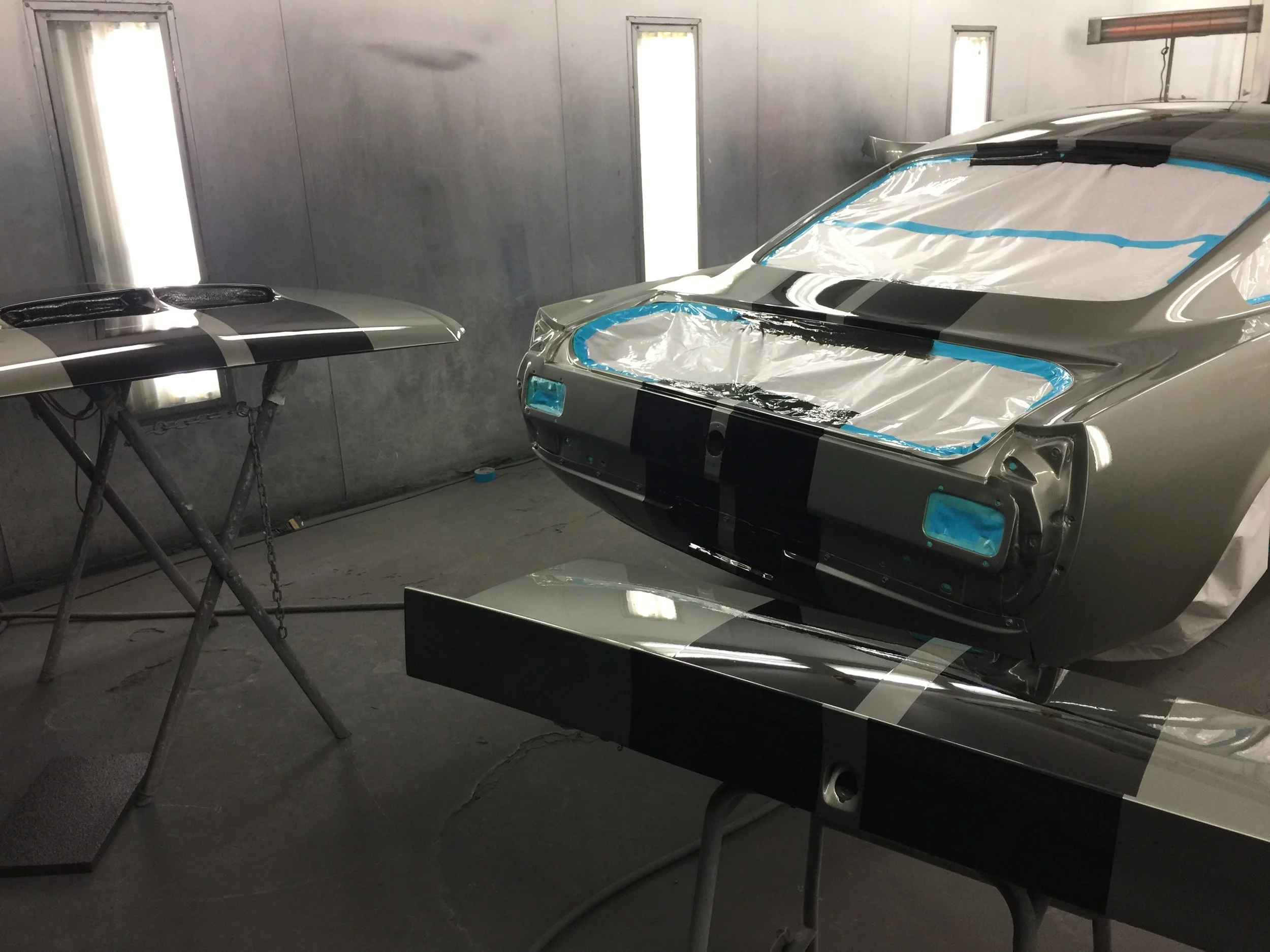 A silver race car is being painted inside a spray booth. The car has taped-off windows, and there is a black and silver spoiler on a separate stand next to it.