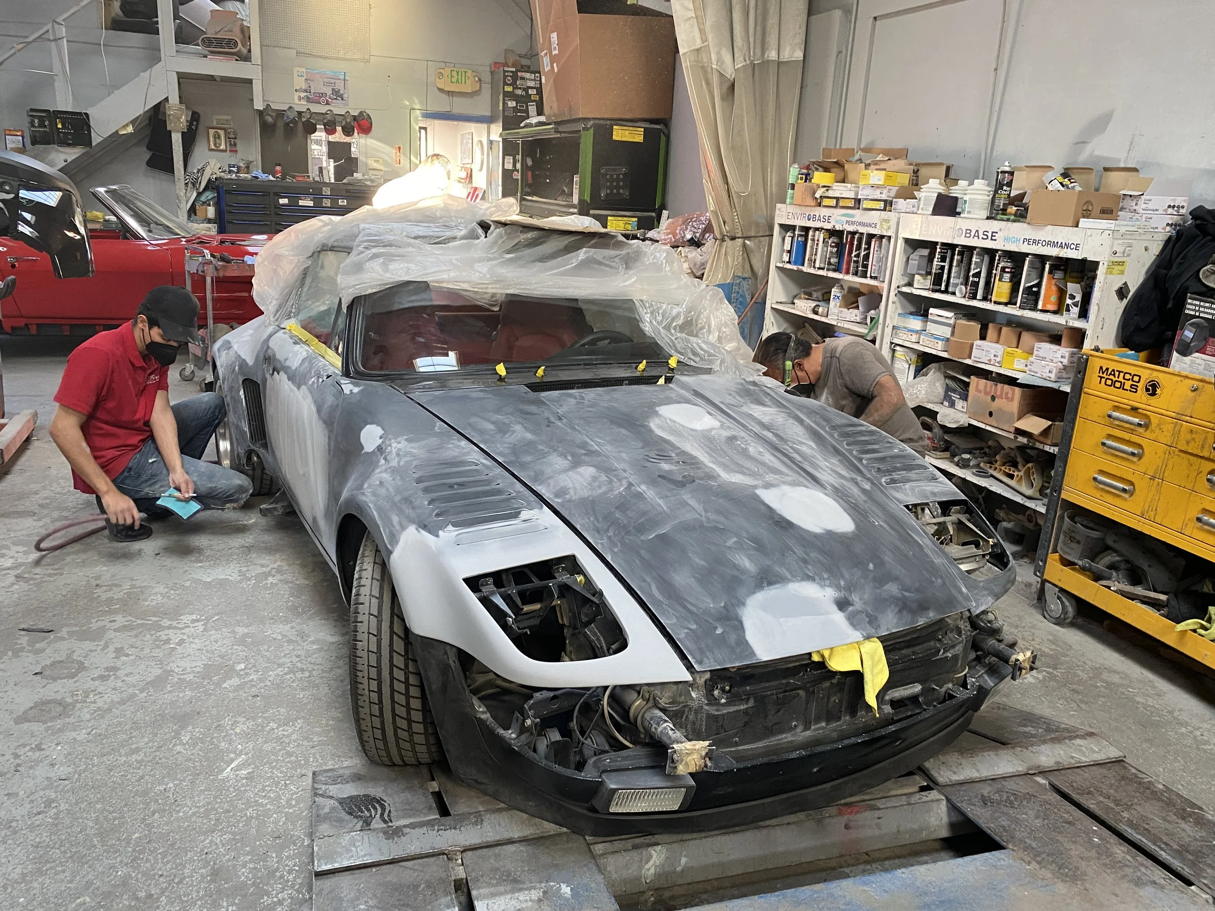 A garage workshop with two workers repairing or restoring a silver sports car. The car is partially painted and covered with plastic. The garage is stocked with tools, shelves of supplies, and a red car in the background.