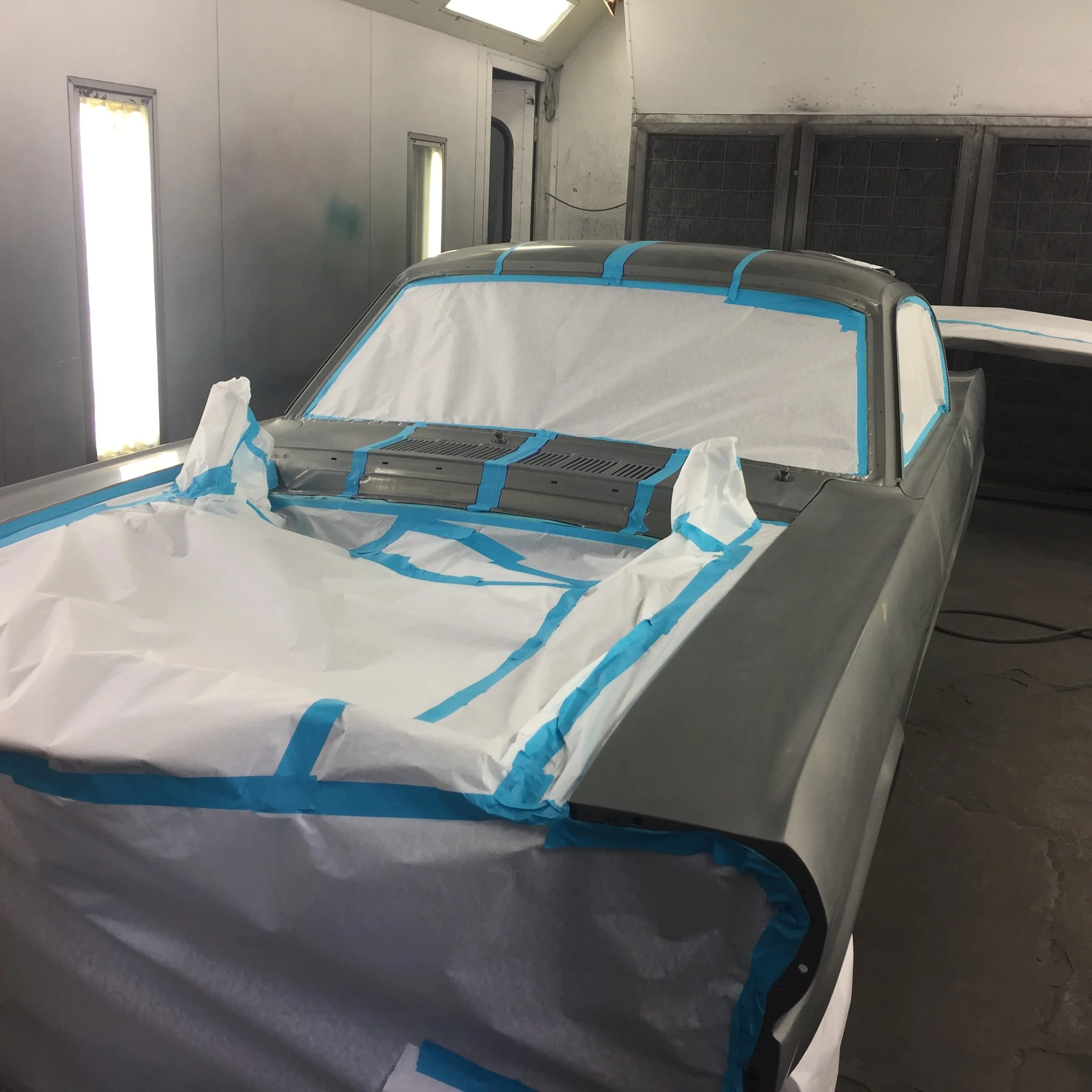 A car in a paint booth, partially masked with blue painter's tape and white paper, preparing for painting or repainting. The setting appears to be an industrial or vehicle workshop.