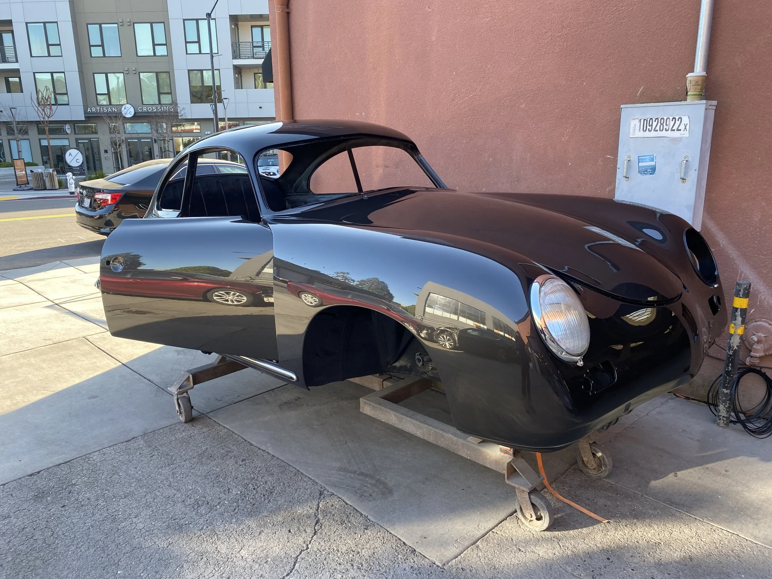 Black vintage car is on a wooden cart outside near a modern building, with a pink wall in the background.