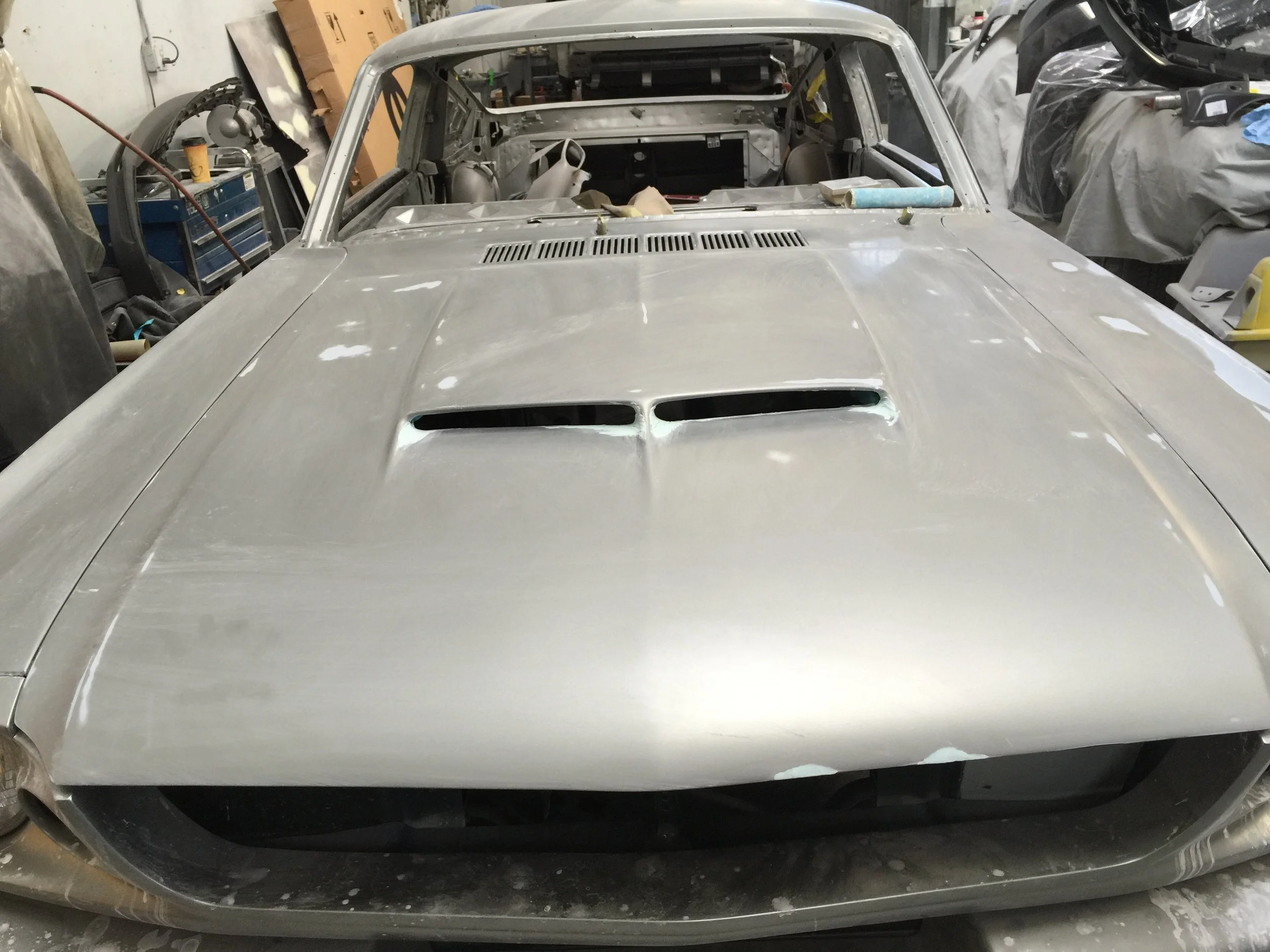 A vintage car body that is under restoration in a workshop, without paint and with visible primer, surrounded by tools and equipment.